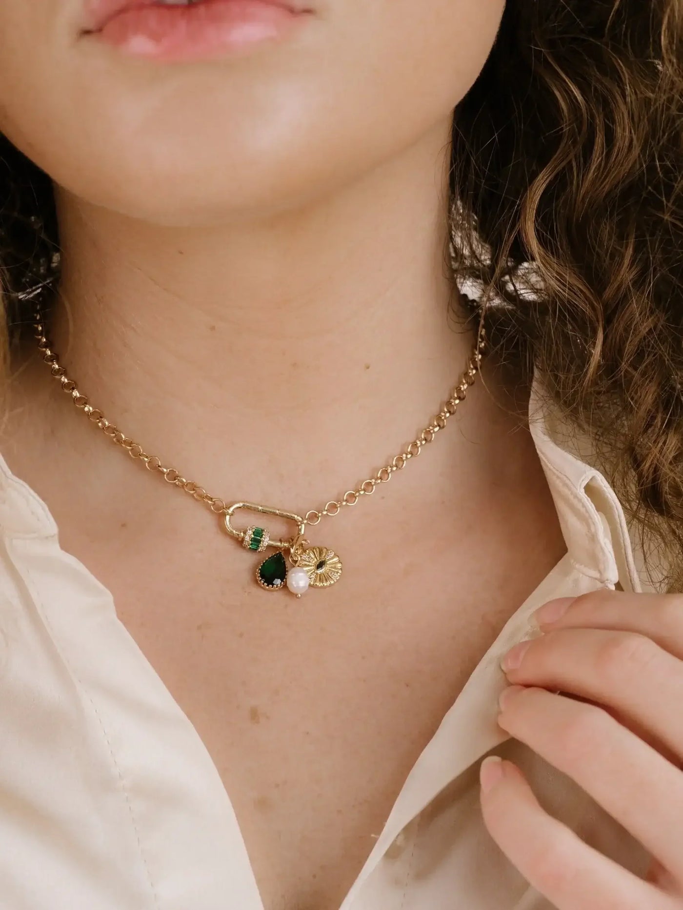 A close-up of a person's neck features a gold chain necklace adorned with green and pearl pendants. The individual is adjusting their shirt collar with one hand in a neutral setting.