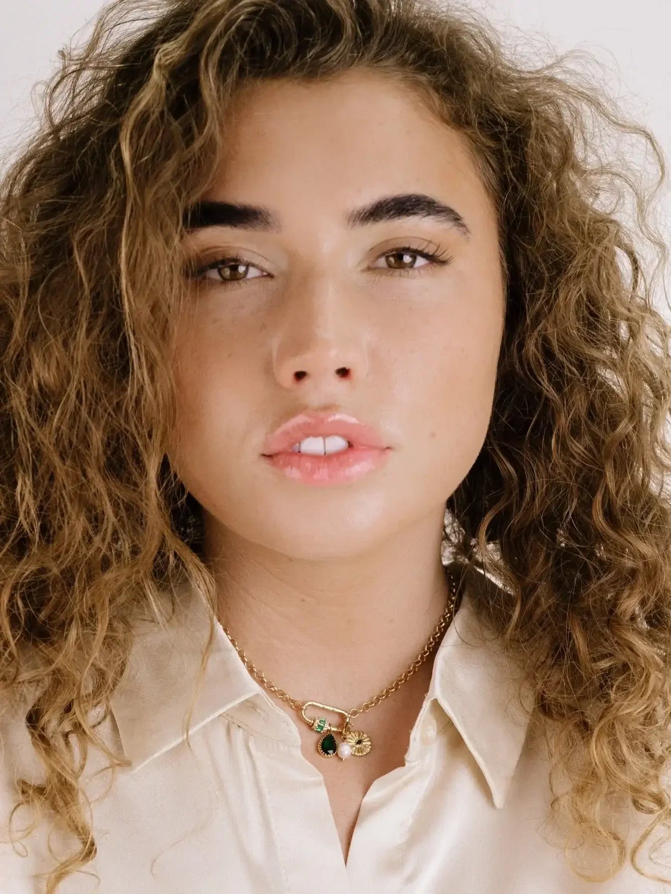 A young woman with curly hair wears a light-colored, button-up shirt and a delicate gold necklace with various gemstones, looking directly at the viewer against a neutral background.