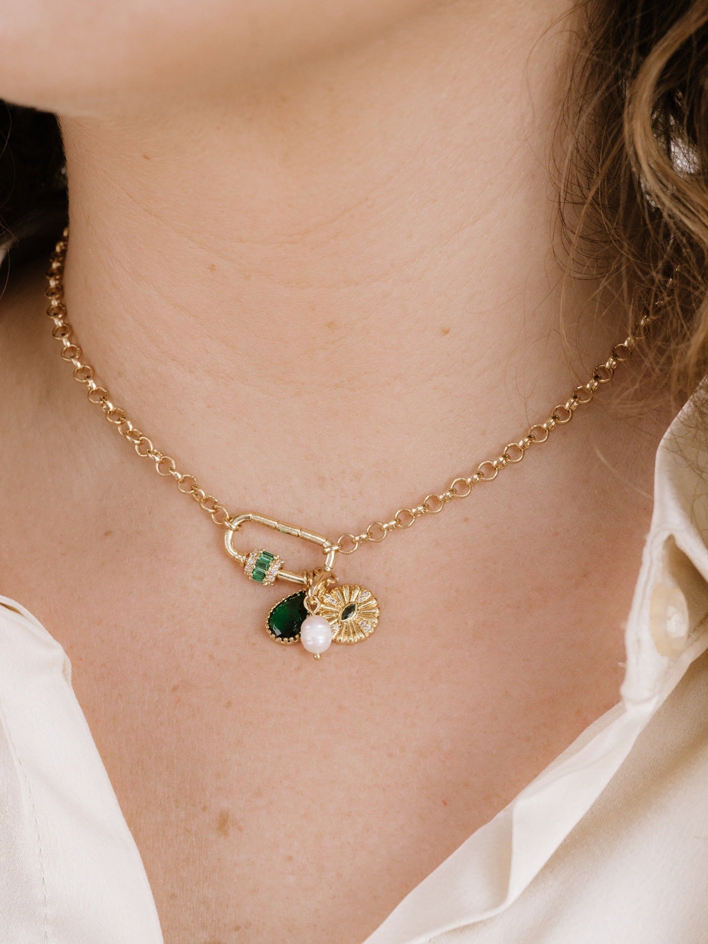 A gold chain necklace adorns the neck, featuring decorative charms like a green gem, a pearl, and a sunflower design. The wearer has light skin and wavy hair.