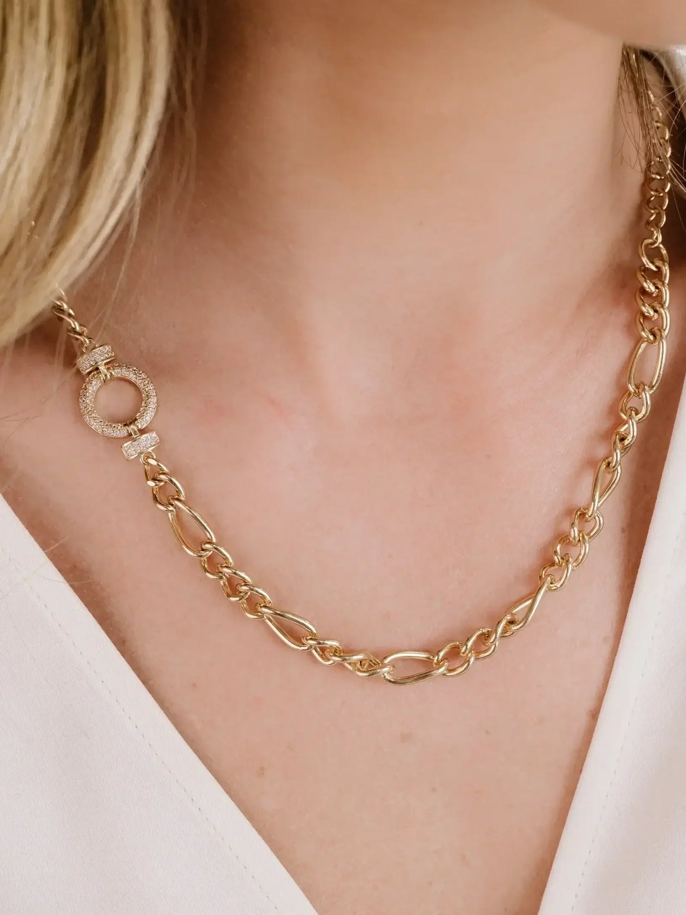A gold chain necklace with interlinked oval and round links, featuring a decorative clasp adorned with sparkling elements, rests elegantly on a person’s neck, against a light-colored top.