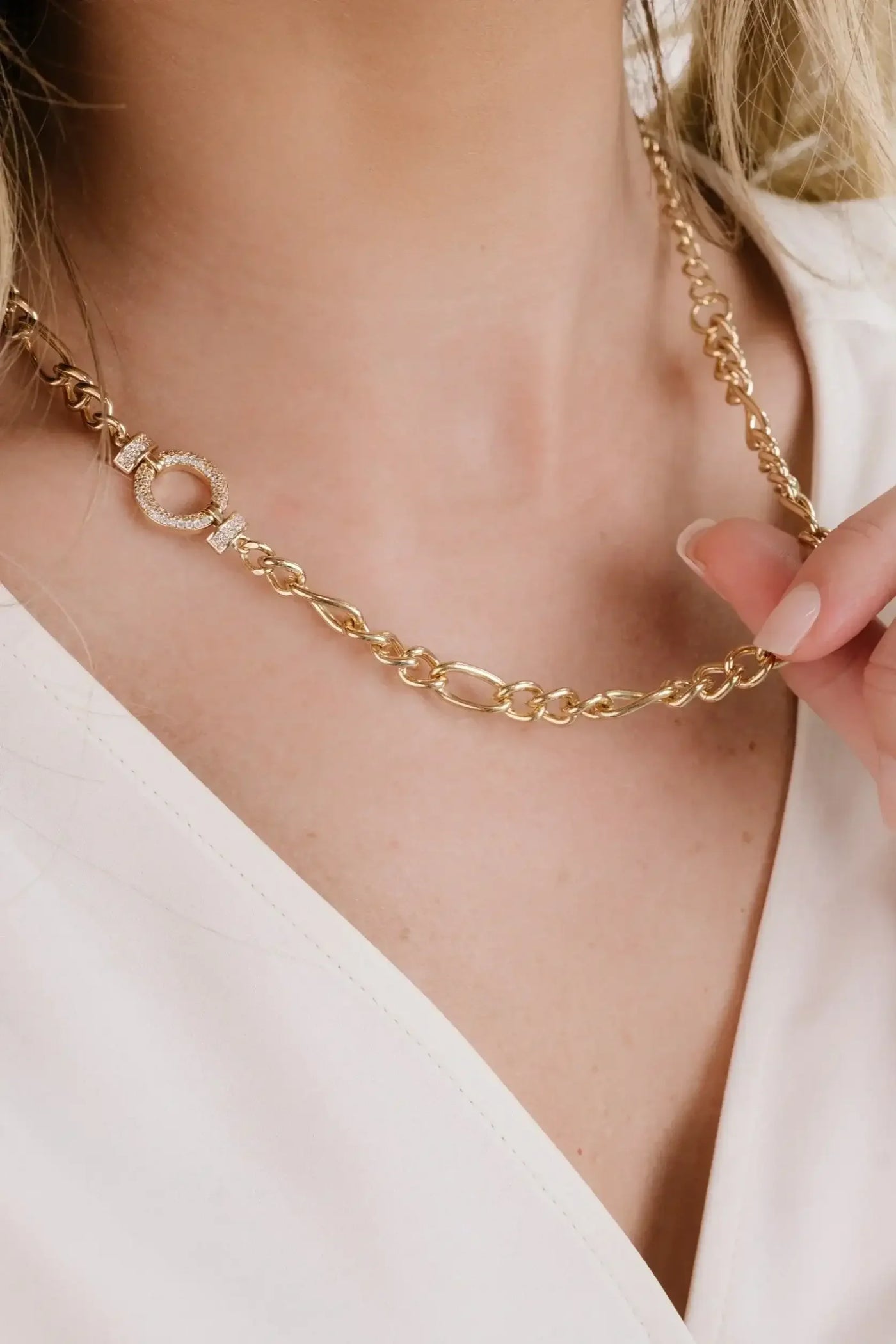 A gold chain necklace with a decorative clasp rests against the skin of a person's neck. The individual gently holds the necklace with manicured fingers, wearing a white top.