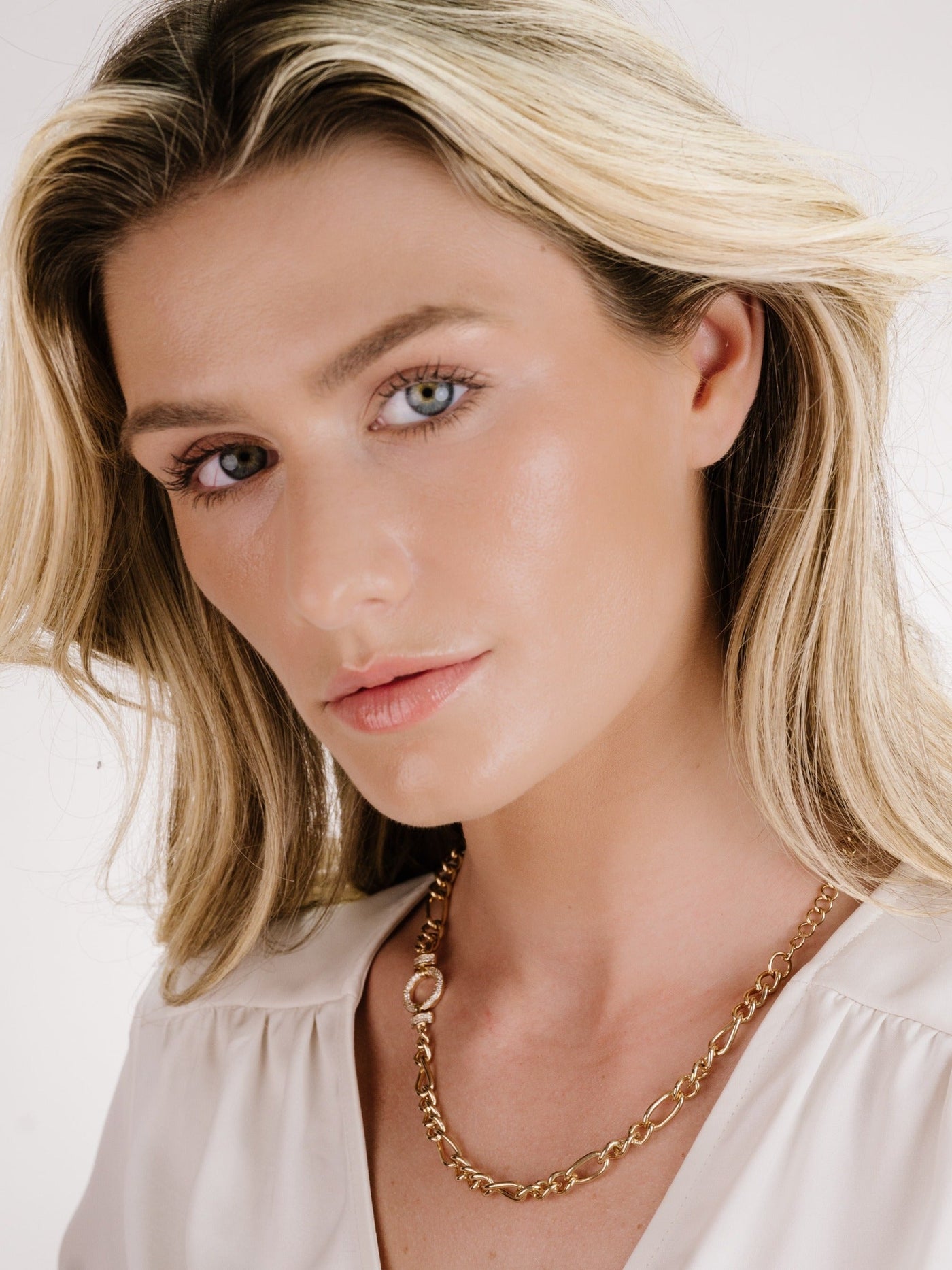 A woman with long, wavy blonde hair poses while wearing a gold chain necklace. She has a natural makeup look, with soft lighting against a neutral background.