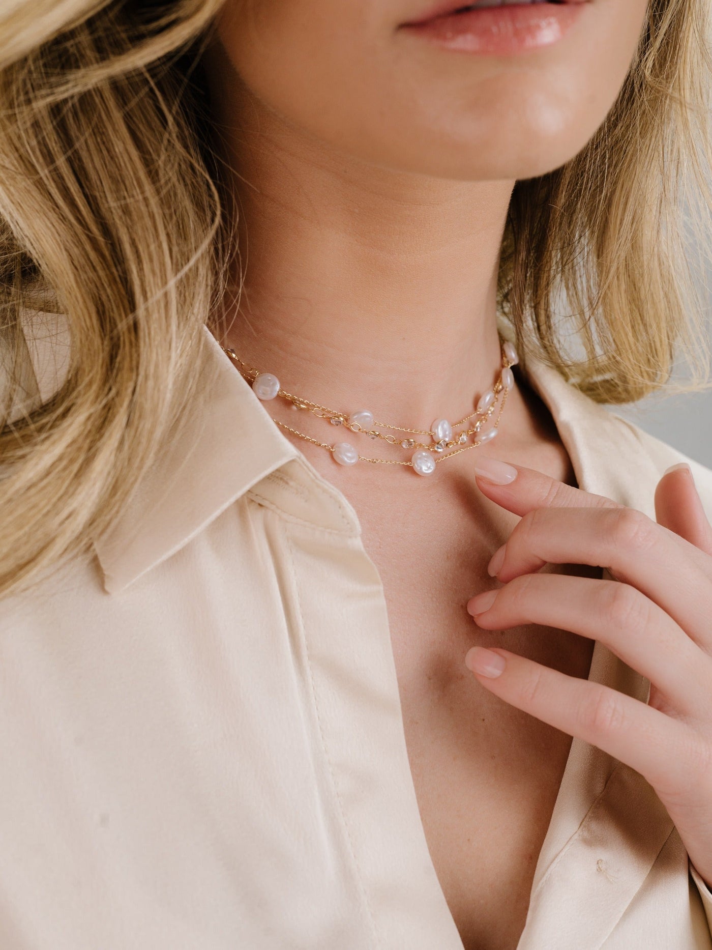 A close-up of a person wearing a delicate layered necklace with pearls, gently touching their neck. They are dressed in a light-colored blouse, with a soft, neutral background.
