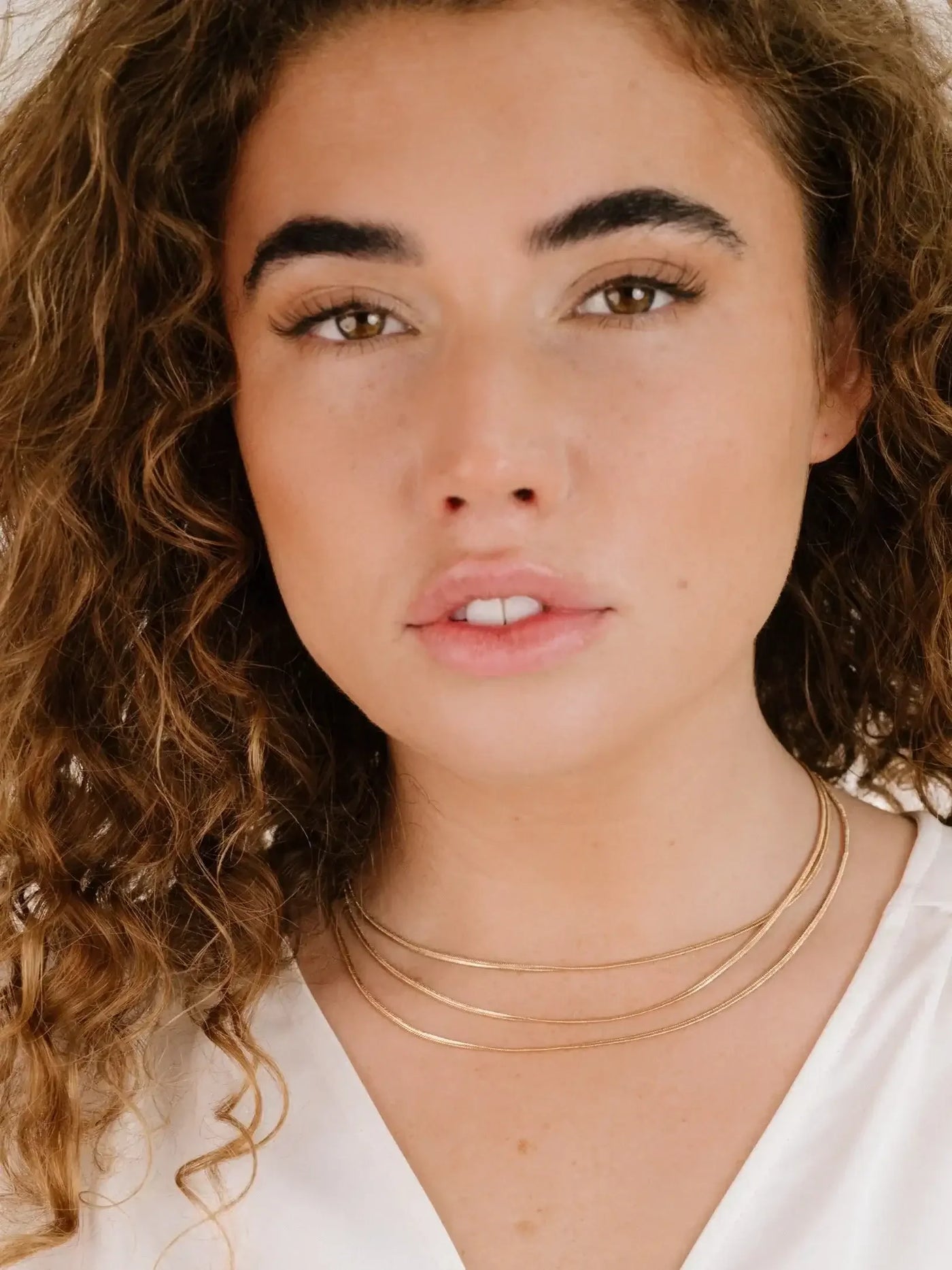 A woman with curly hair is looking directly at the camera, wearing a white top and layered gold necklaces. Her expression is neutral, set against a light background.