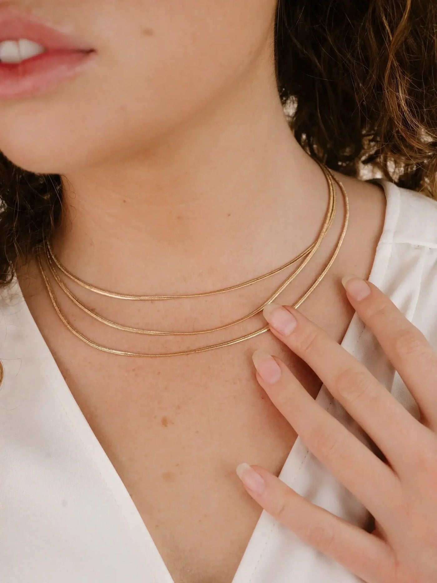 A person is wearing multiple gold necklaces, gently touching one with their right hand. The setting features a close-up view of their neck and upper chest, displaying smooth skin.