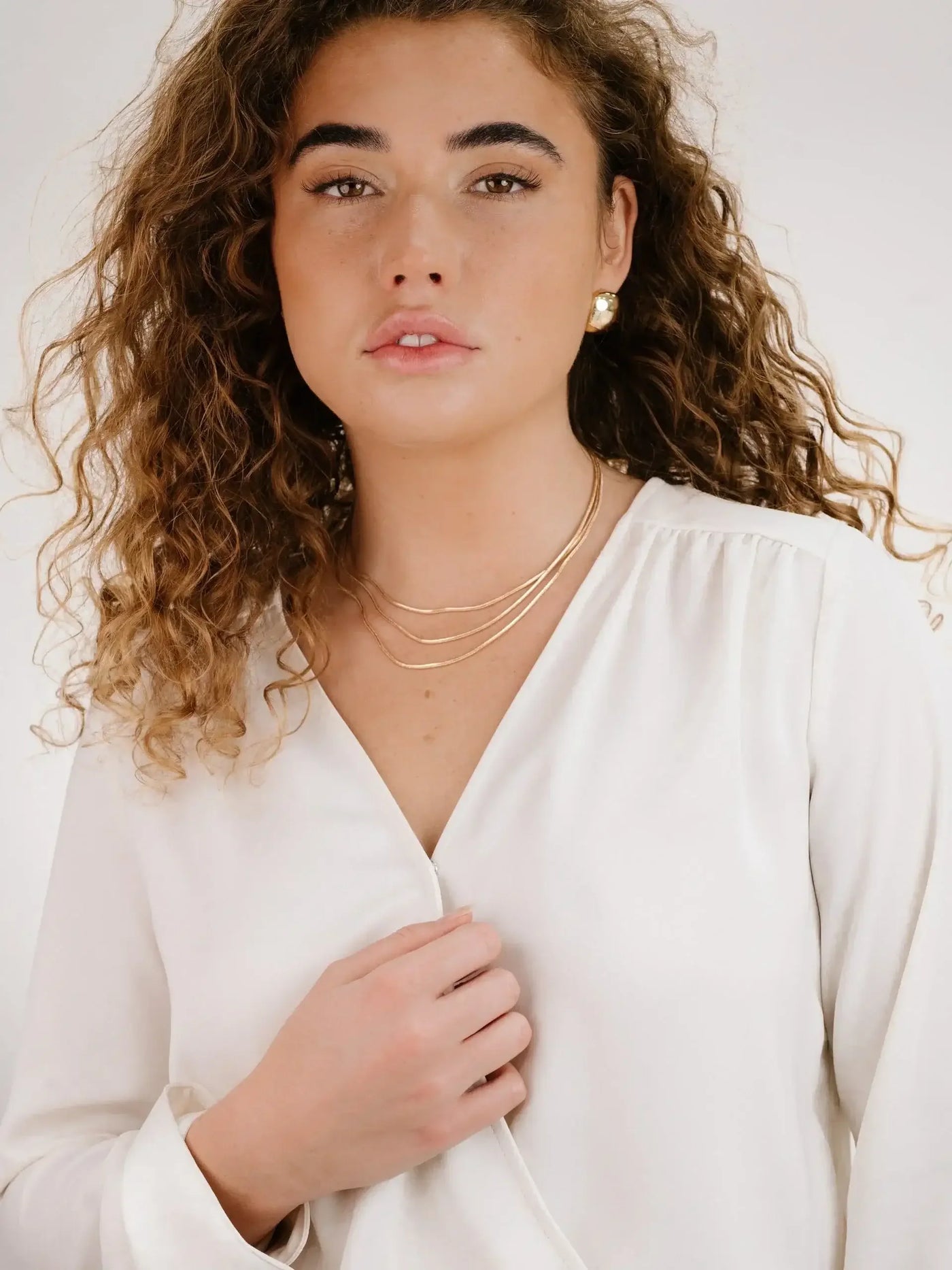 A woman with curly hair wears a white blouse and layered gold necklaces, looking directly at the camera. Her hand rests lightly on her chest, conveying a confident expression against a plain background.