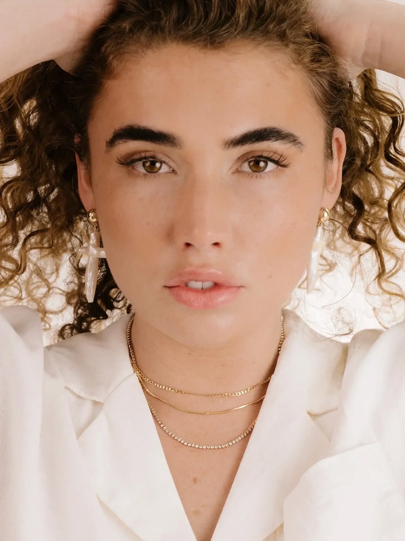 A woman with curly hair poses confidently, touching her hair. She wears layered necklaces and large earrings, against a simple, light background that enhances her striking features.