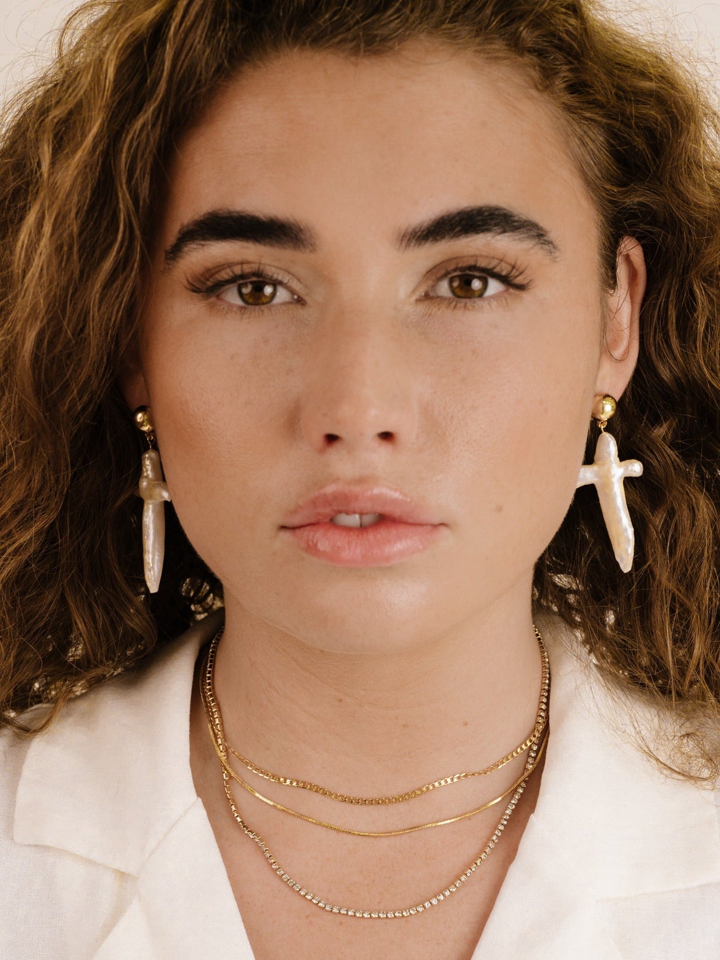 A young woman with curly hair wears elegant, dangling earrings and layered gold necklaces. She poses against a neutral background, exuding confidence with a subtle expression on her face.