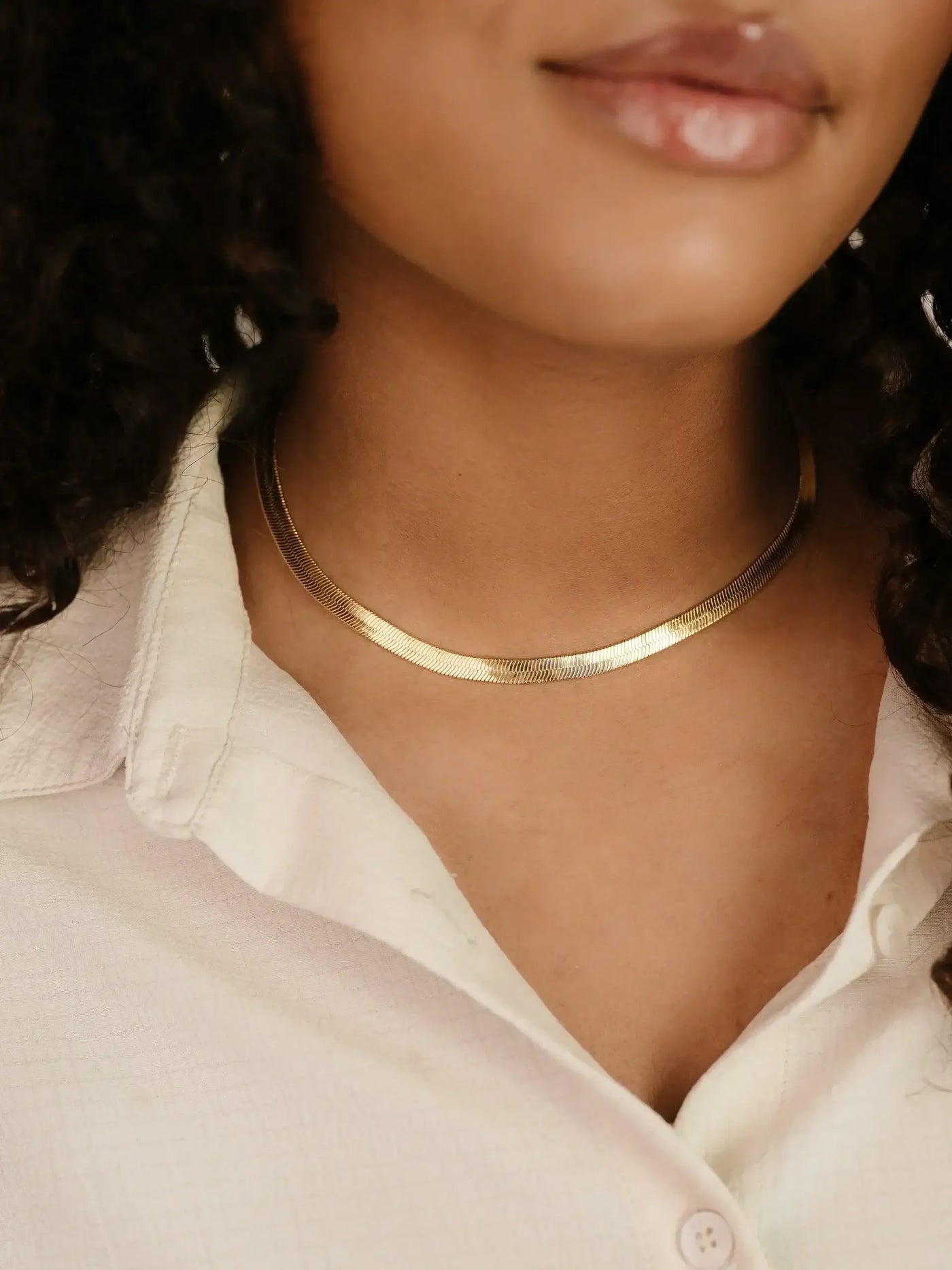 A gold choker necklace rests gracefully on a woman's neck. She wears a light-colored, textured shirt, with her wavy hair partially visible, set against a soft background.