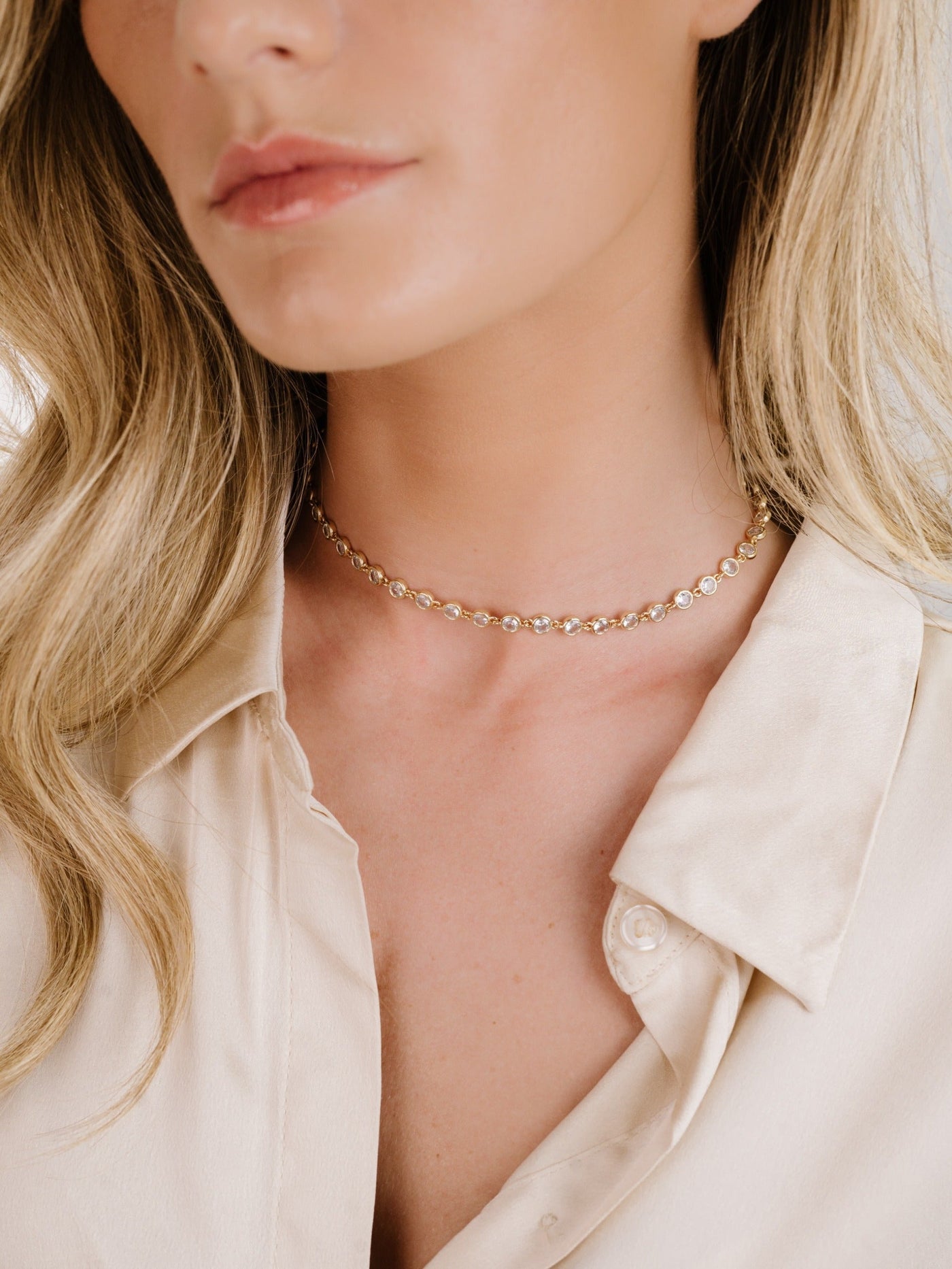 A close-up of a woman’s neck adorned with a delicate gold choker featuring circular embellishments, set against a softly lit background. The woman wears a light-colored, silky shirt.