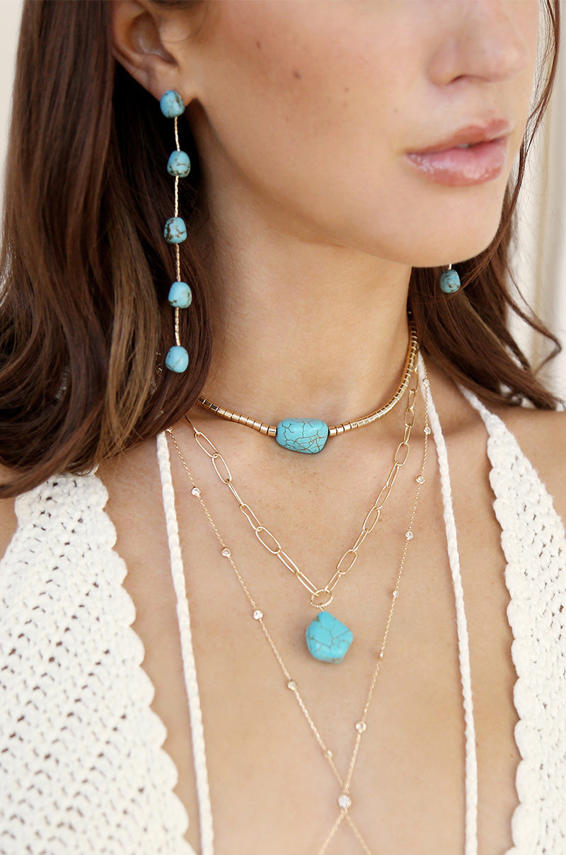 A woman wears multiple layered necklaces featuring turquoise stones and gold accents, with coordinating turquoise earrings. The setting appears light and casual, highlighting a bohemian style.