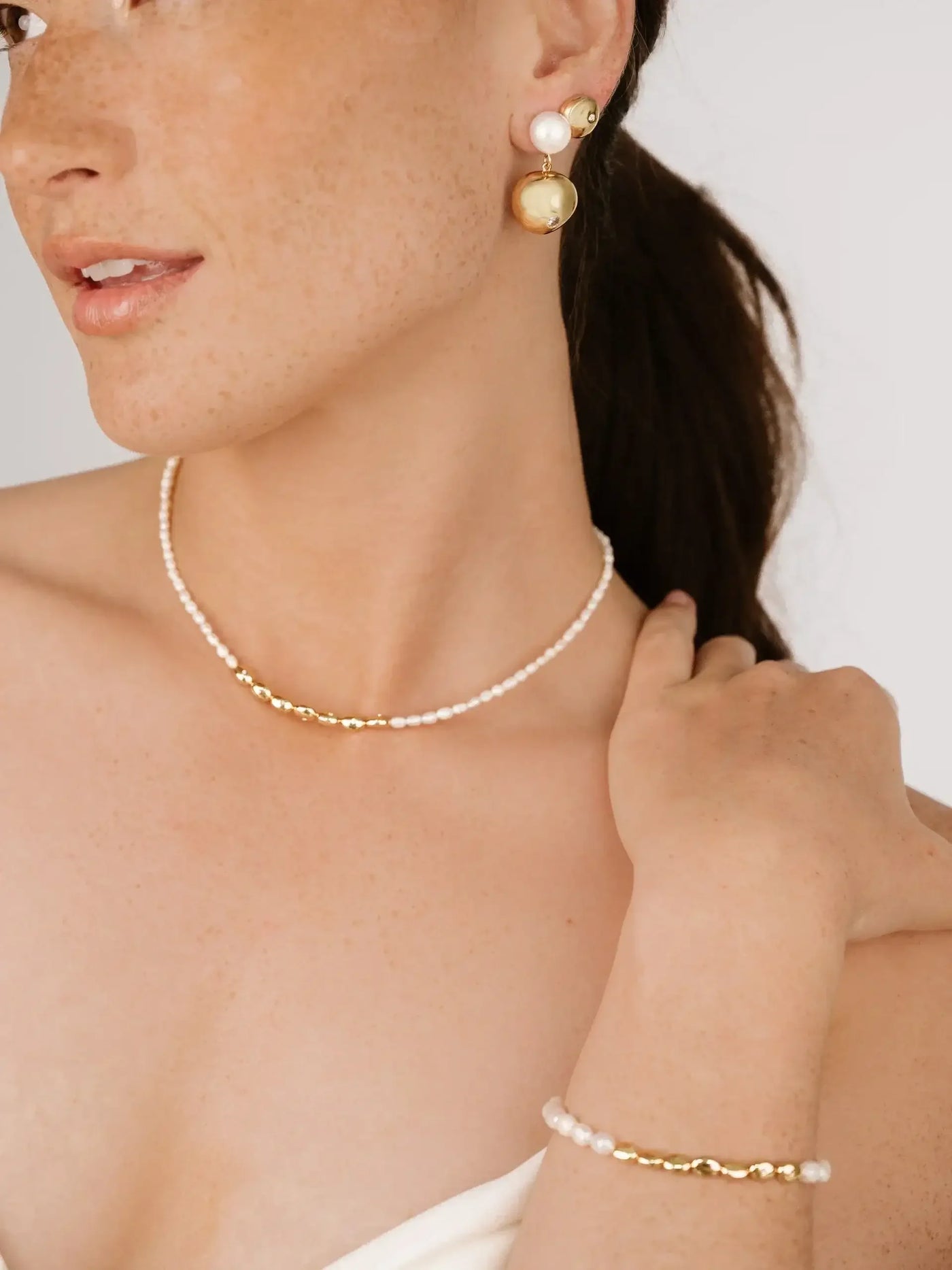 A woman with freckles showcases elegant jewelry, including a pearl necklace and earrings, while gently touching her shoulder, set against a neutral background.