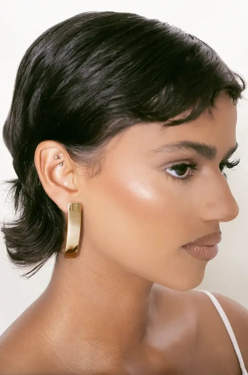 A woman with short, wavy hair is wearing large, shiny gold hoop earrings. She has a natural makeup look, including a highlighted cheekbone, against a soft, neutral background.