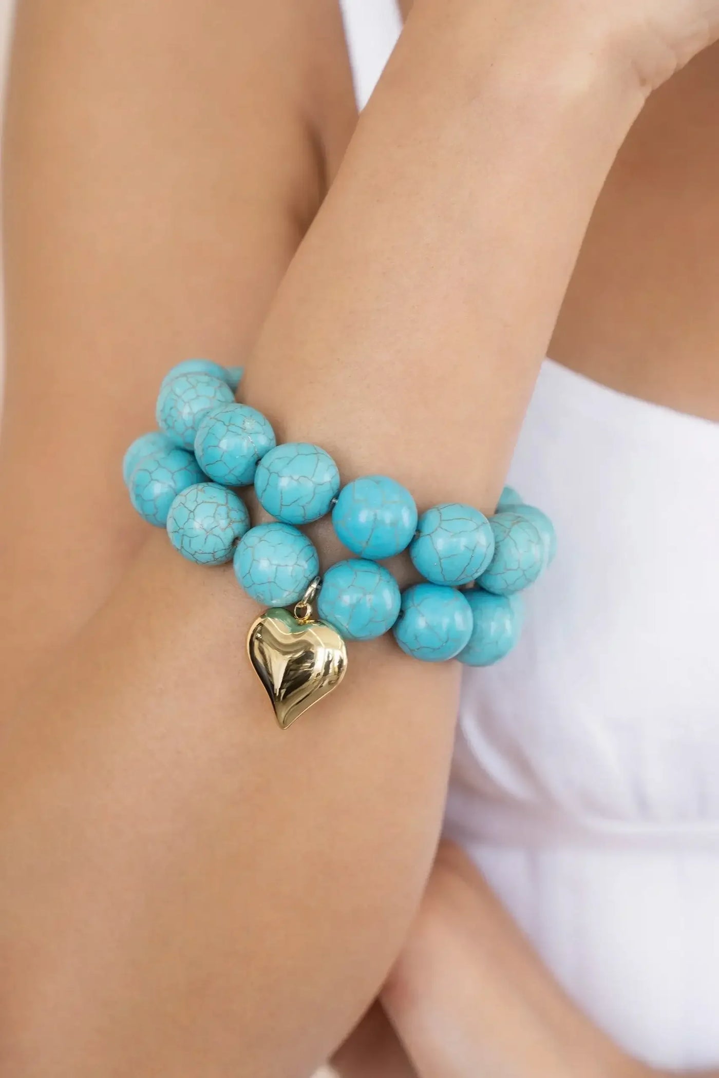 A turquoise beaded bracelet adorned with a golden heart charm embraces the wrist of a person in a white top, showcasing the accessory against a neutral background.