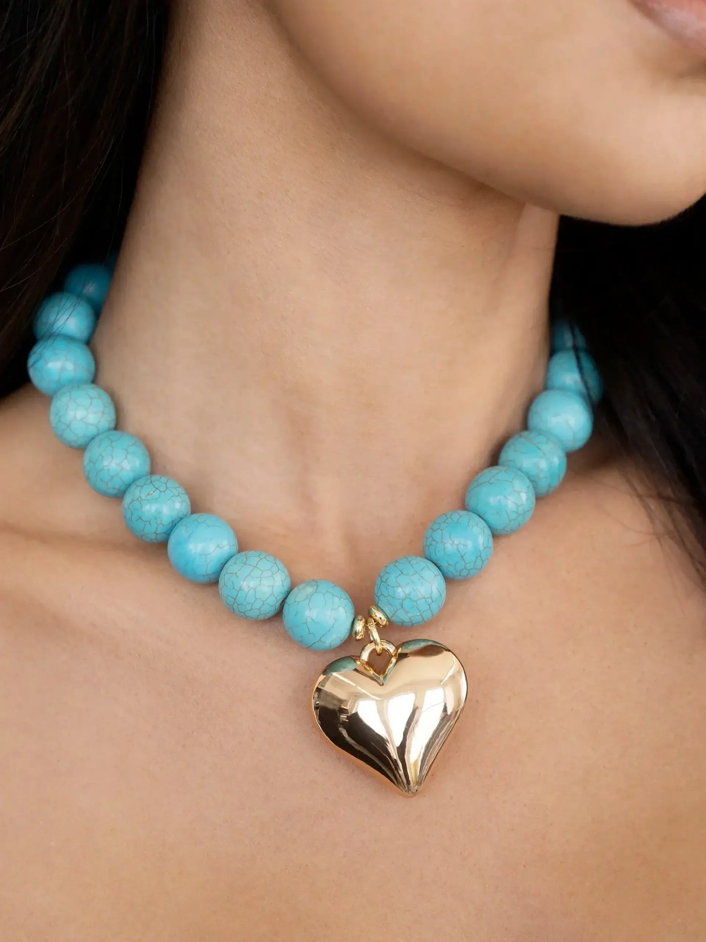 A turquoise beaded necklace adorns a neck, featuring a prominent golden heart pendant. The setting is neutral, focusing on the jewelry's color contrast against skin tone.