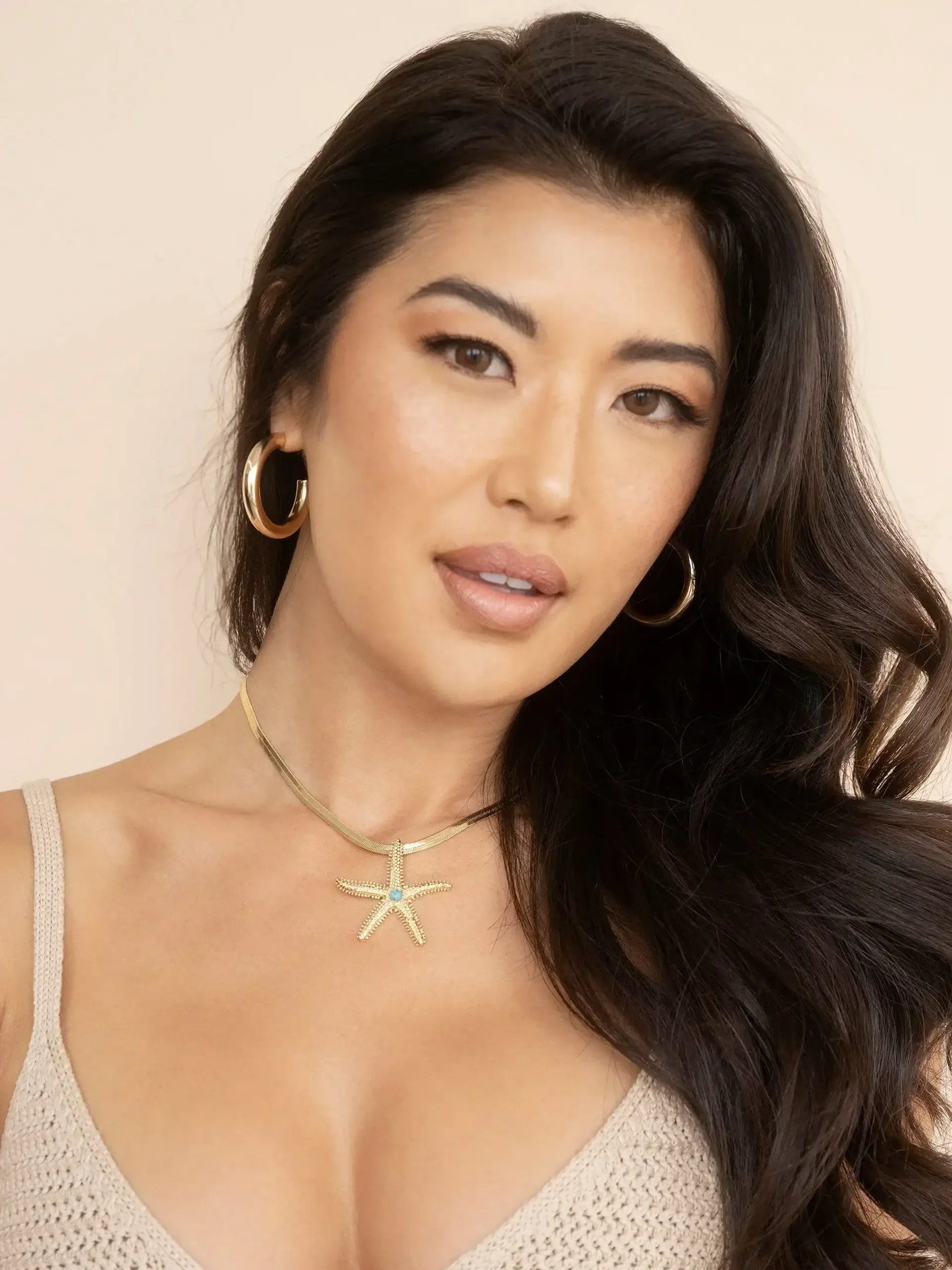 A woman with long, dark hair wears a gold starfish pendant necklace and hoop earrings. She is posed against a soft beige background, displaying a confident expression.