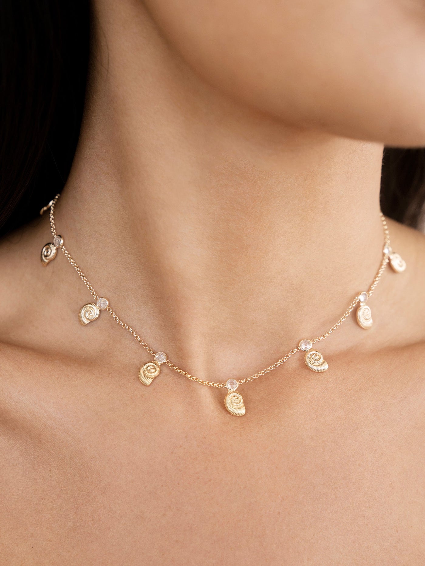 A delicate gold necklace with spiral shell motifs and small beads hangs around a woman's neck, accentuating her skin in a softly lit, neutral environment.