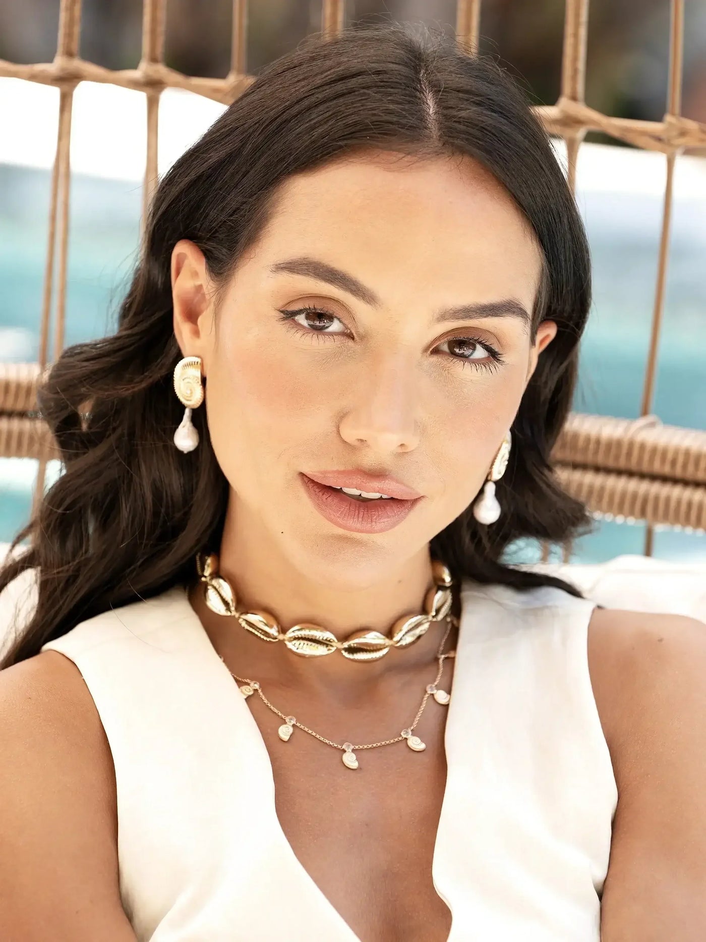 A woman with long, dark hair sits relaxed, wearing multiple golden necklaces and distinctive shell-shaped earrings. Soft outdoor lighting suggests a tranquil, stylish atmosphere, with a blurred pool in the background.