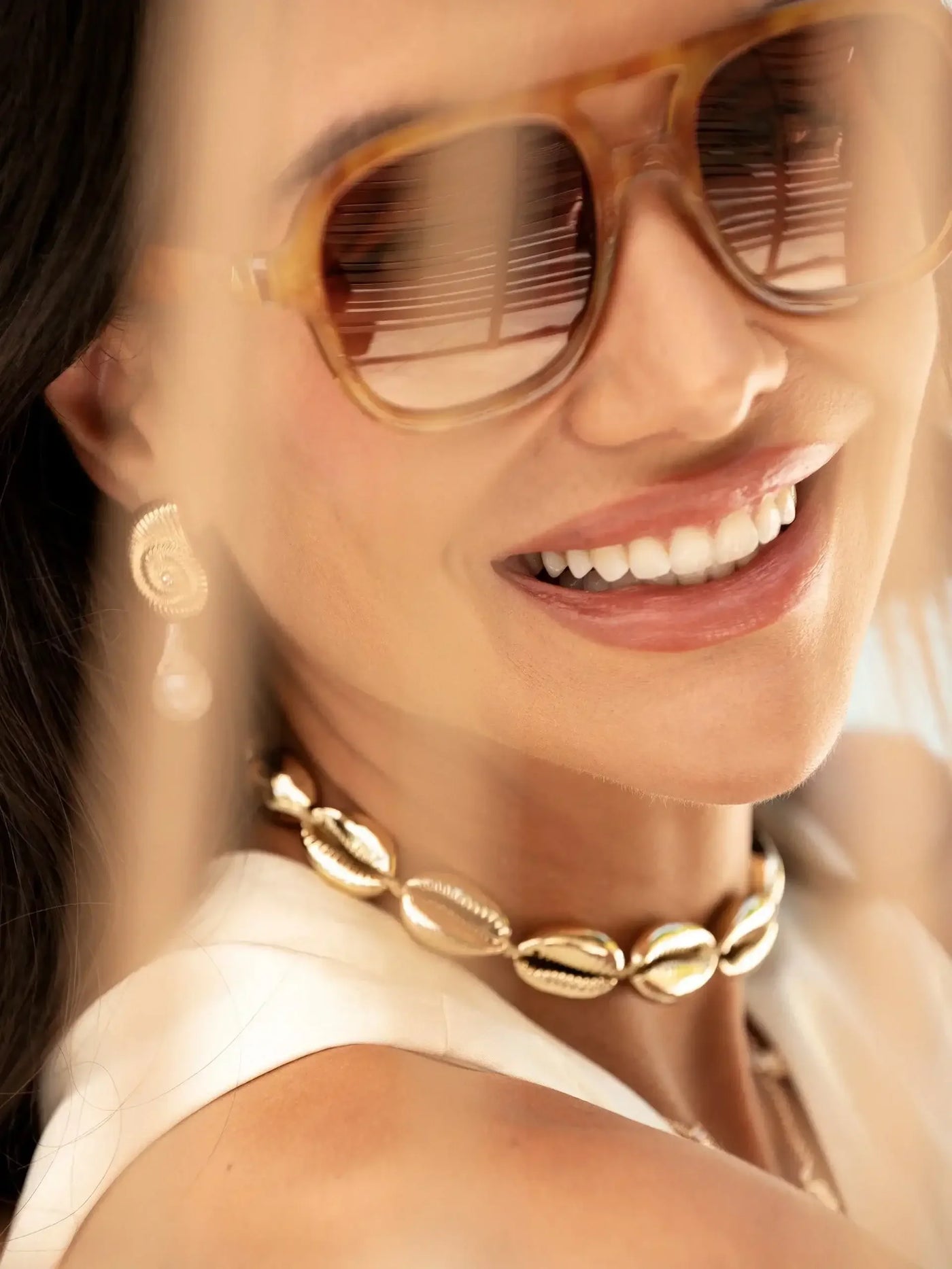 A woman smiles brightly, wearing stylish sunglasses and a gold shell necklace. She appears relaxed, with soft light illuminating her face, suggesting a warm, sunny environment.