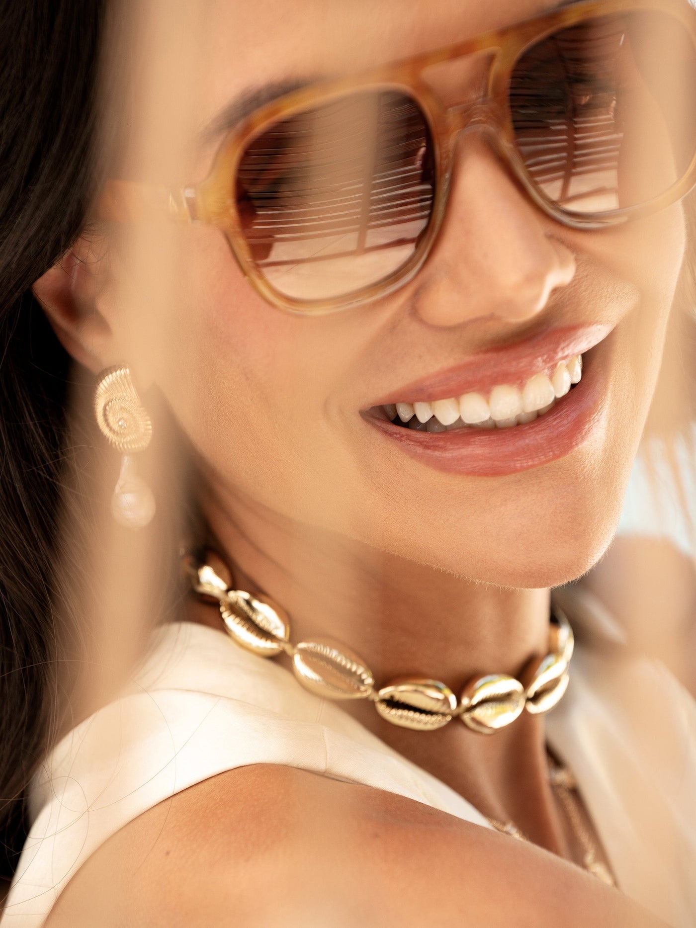 A woman smiles brightly, wearing stylish sunglasses and a gold shell necklace. She appears relaxed, with soft light illuminating her face, suggesting a warm, sunny environment.