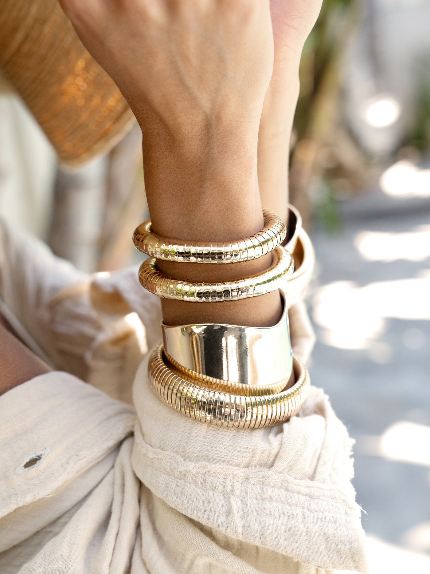 Multiple shiny gold bangles adorn a person's wrist, with a mix of smooth and textured designs. The background is softly blurred, featuring hints of greenery and subtle light reflections.