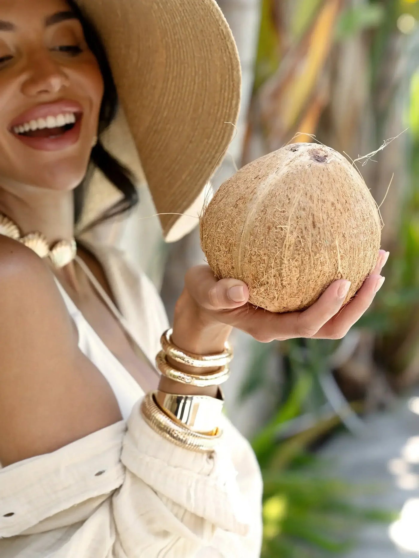 A woman joyfully holds a coconut, wearing a wide-brimmed hat and multiple gold bracelets. She stands amidst tropical greenery, exuding a relaxed, sunny vibe.