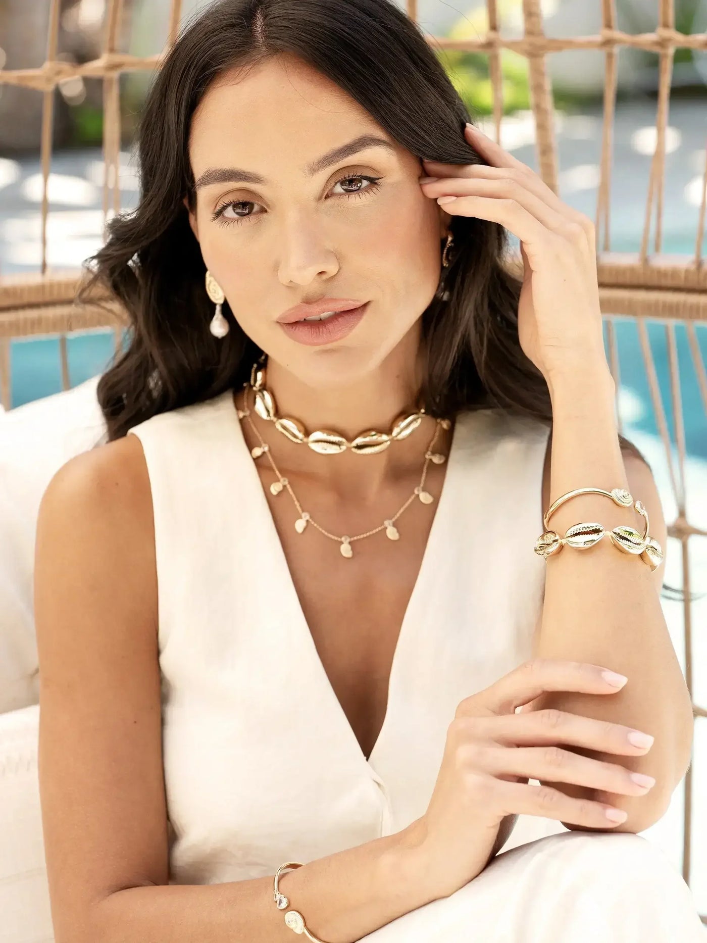 A woman with long, dark hair touches her face while wearing elegant jewelry, including a necklace and bracelets, in a bright, airy outdoor setting with a blurred natural background.
