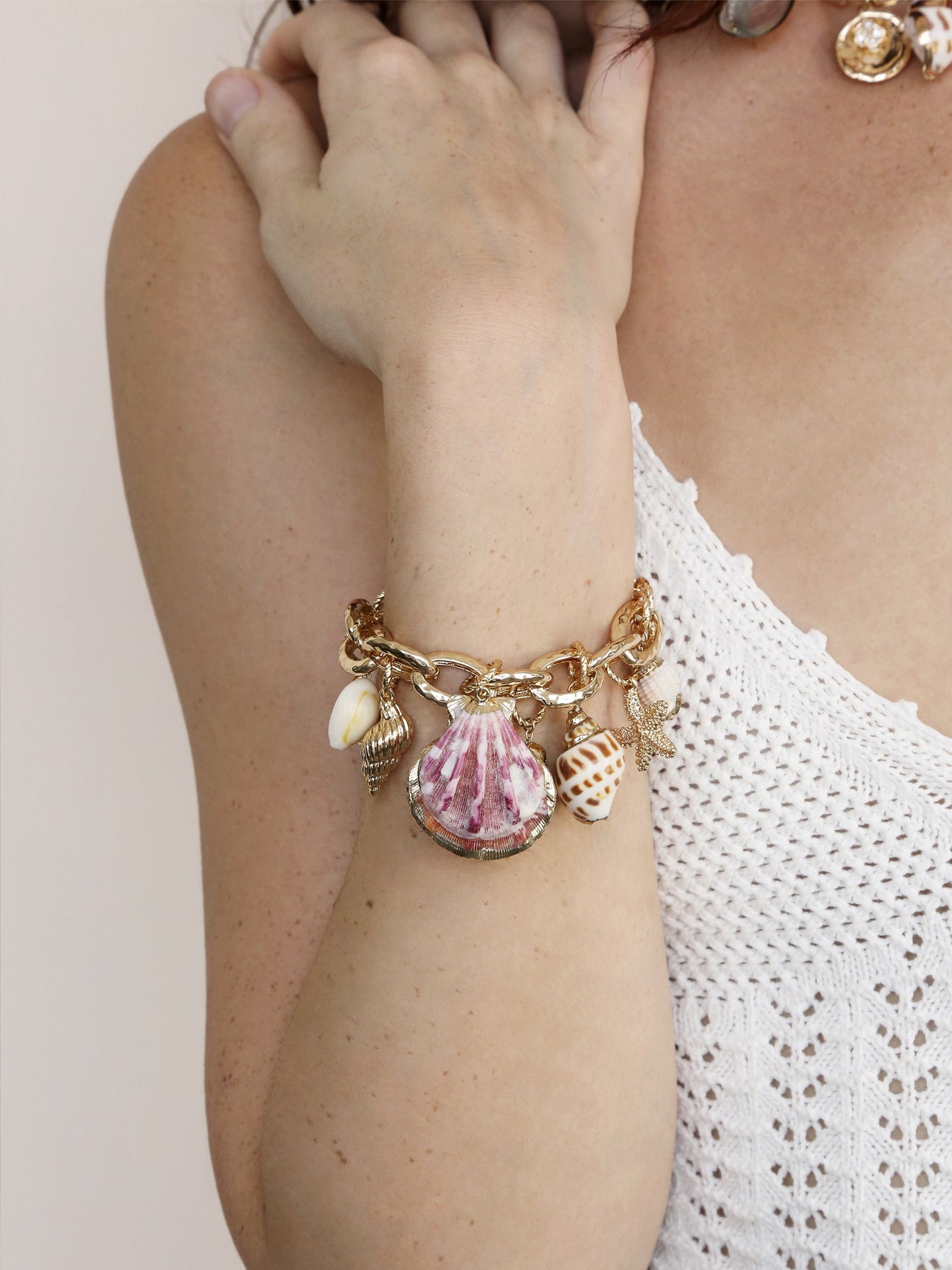 A gold charm bracelet adorned with seashells hangs on a woman's wrist, while she lightly touches her shoulder. The background features a soft, neutral tone.