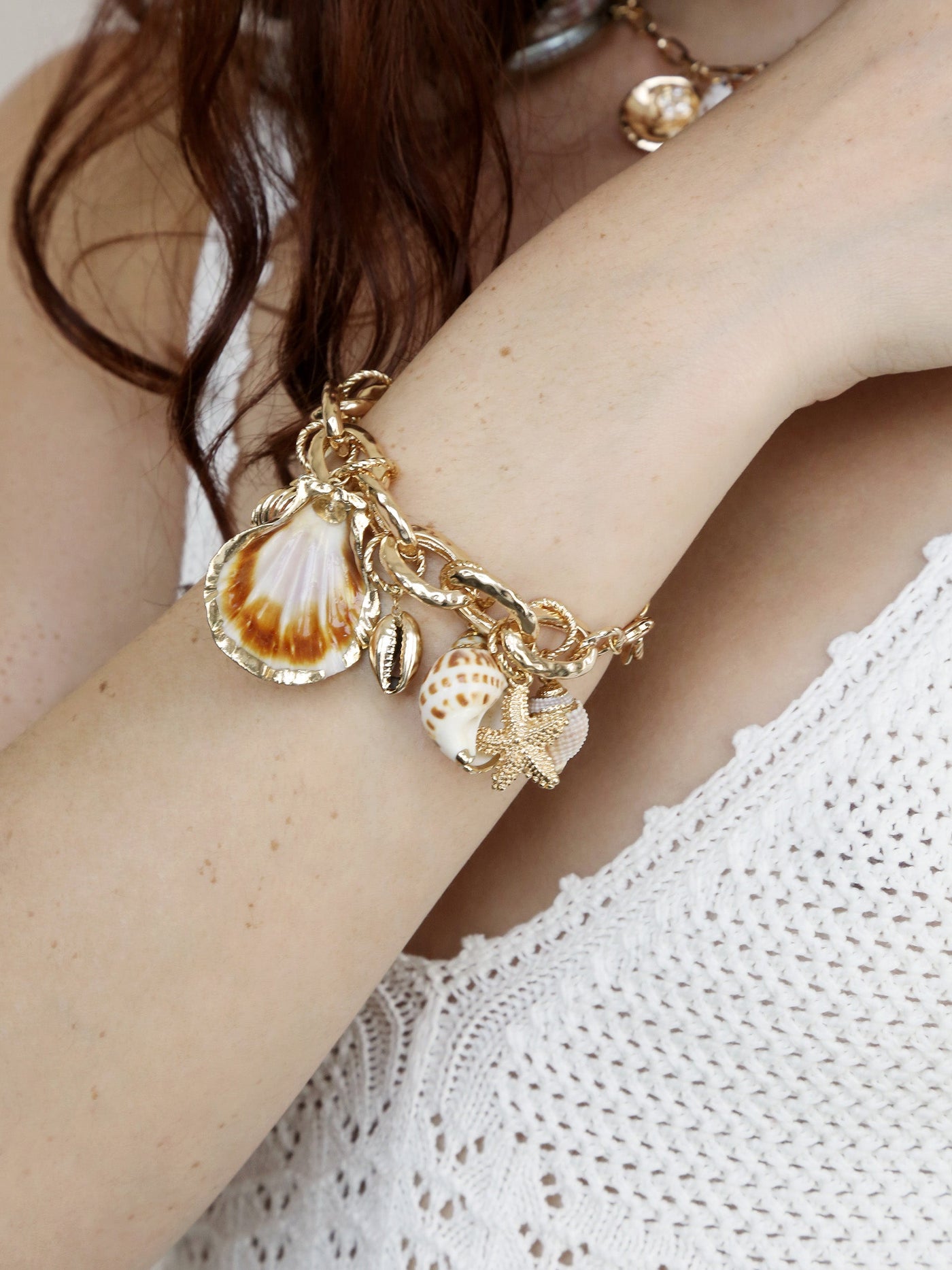 A gold charm bracelet adorned with seashells and a starfish rests on a bare arm. The wrist is partially covered by a knitted white garment, suggesting a casual, coastal aesthetic.