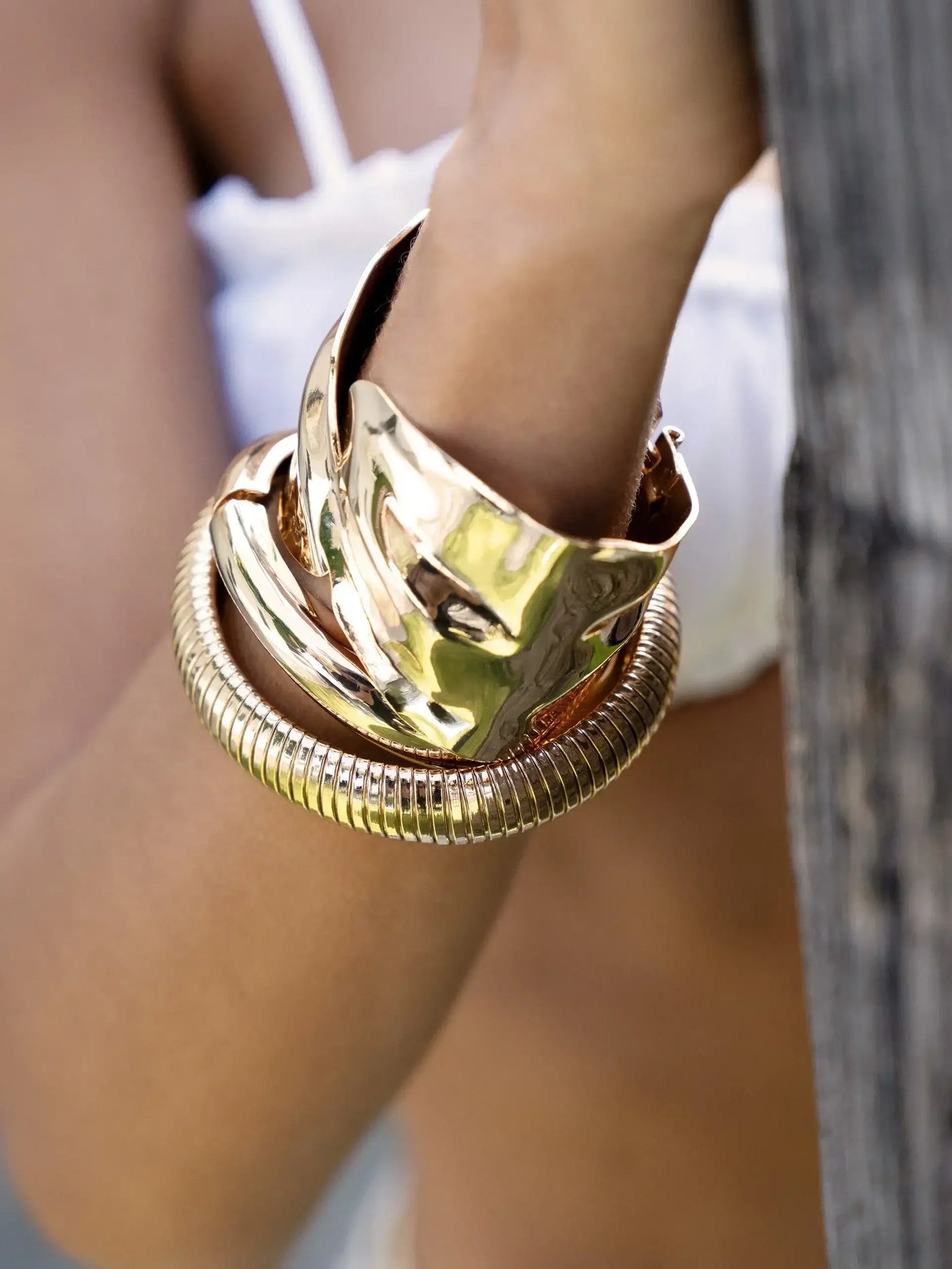 A wrist adorned with three metallic bracelets, including a smooth, wide cuff and a spiral-textured band. The hand rests against a wooden surface, partially blurred background suggests an outdoor setting.