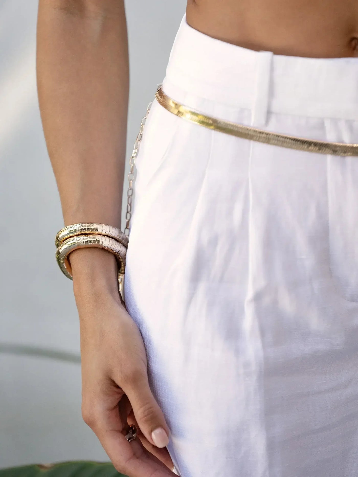 A hand rests on a white high-waisted garment, adorned with a gold belt and layered bracelets. The background is softly blurred, hinting at a natural, outdoor setting.