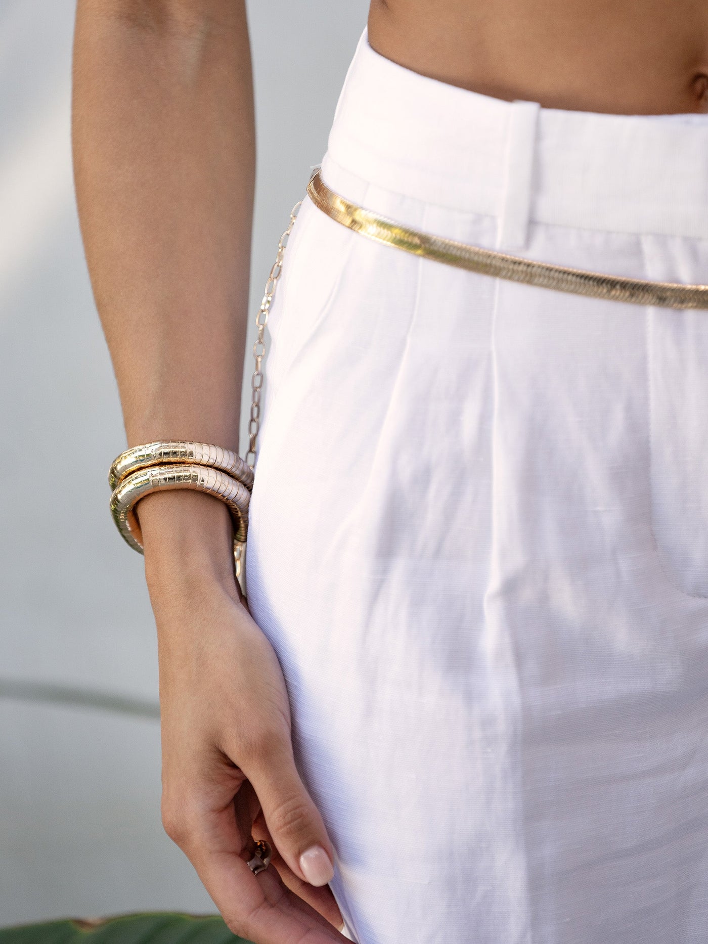 A hand rests on a white high-waisted garment, adorned with a gold belt and layered bracelets. The background is softly blurred, hinting at a natural, outdoor setting.