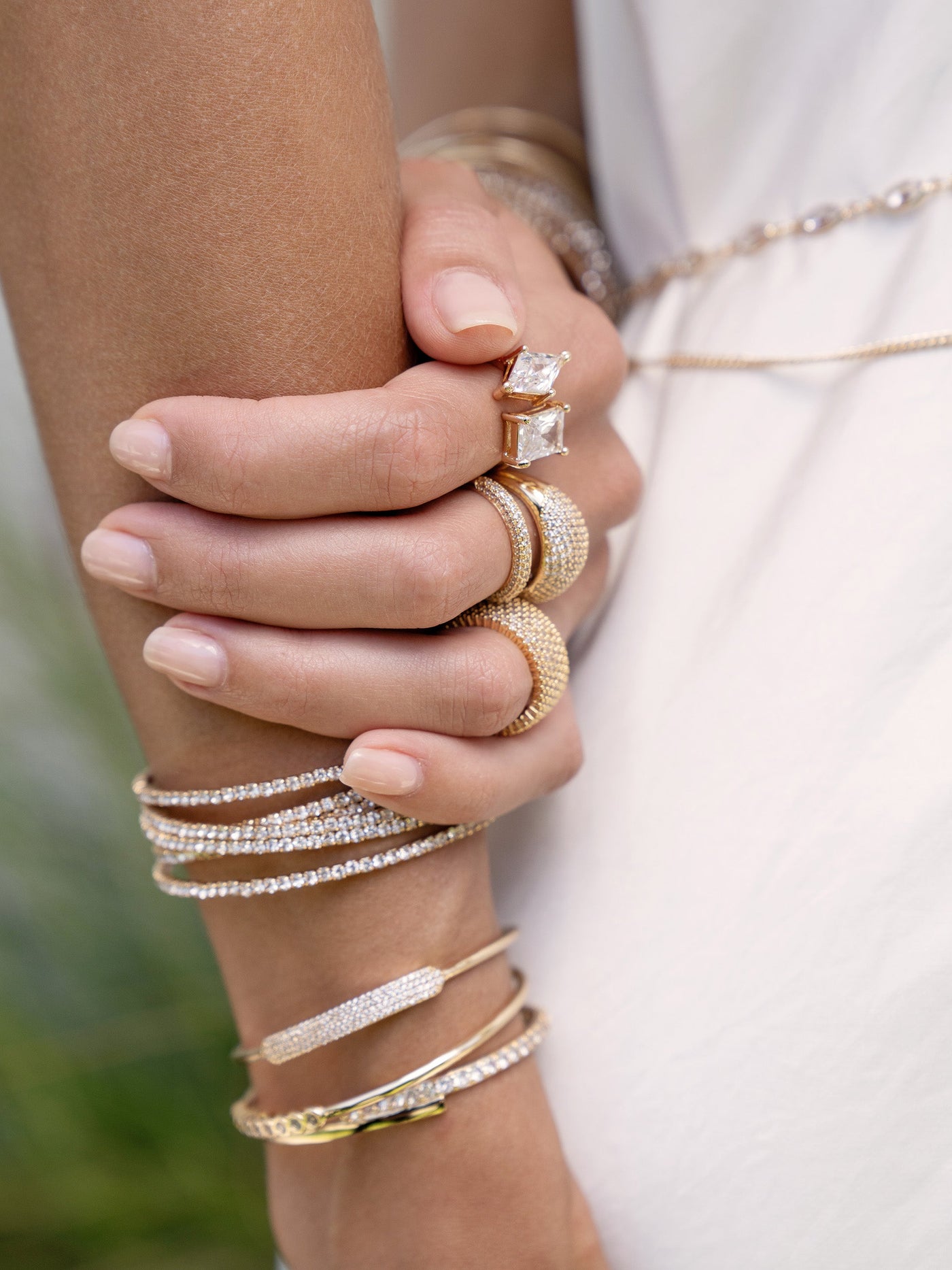A hand adorned with multiple gold and diamond rings and bracelets grips another hand. The setting appears soft and natural, with blurred greenery in the background.