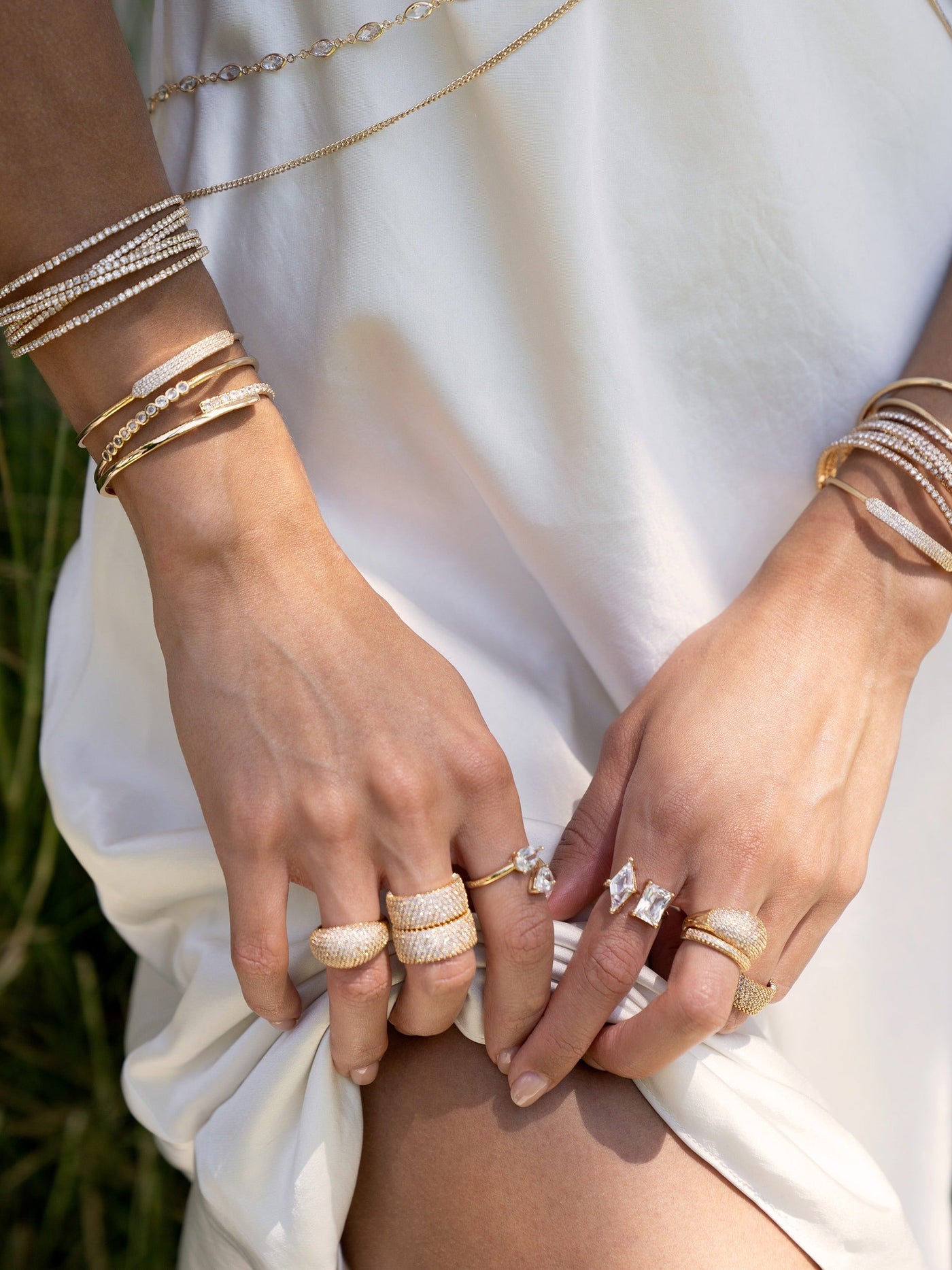 A pair of hands adorned with multiple gold and diamond rings and bracelets grasp the hem of a flowing white dress, set against a backdrop of greenery under bright sunlight.