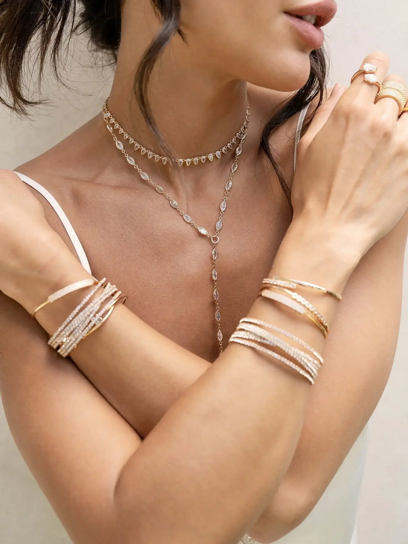 A person displays multiple layered necklaces and intricately designed bracelets on their arms. The soft, neutral background emphasizes the elegance of the jewelry while highlighting the person’s graceful pose.