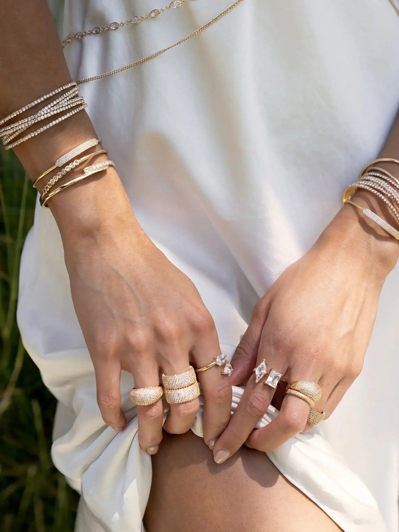 Hands adorned with multiple rings and bracelets are gently holding the edge of a white, flowing garment. The background suggests a natural setting with soft green grass, enhancing the delicate scene.