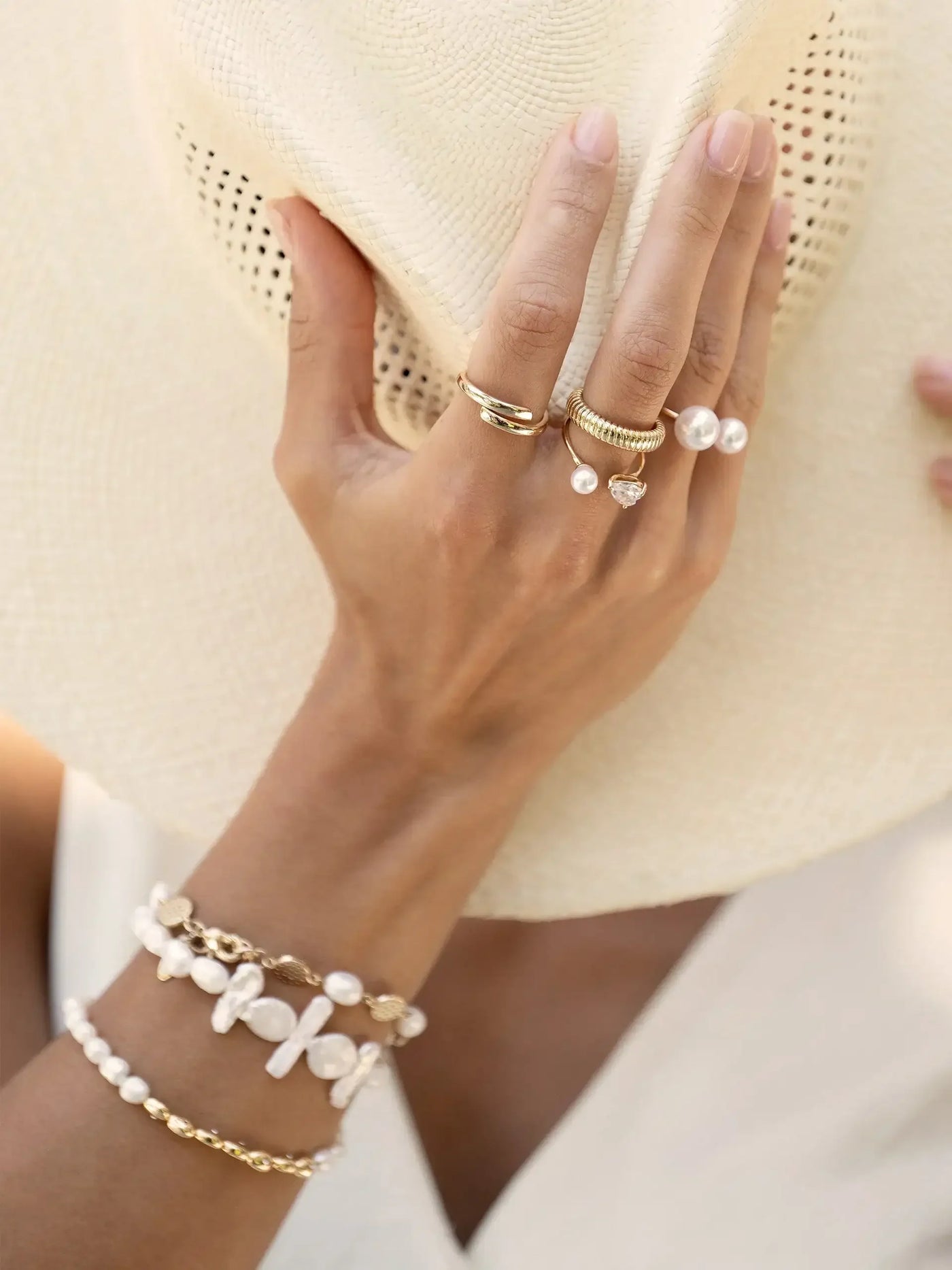 A hand adorned with multiple rings and bracelets elegantly holds a wide-brimmed straw hat, set against a light, neutral background suggesting a sunny outdoor environment.