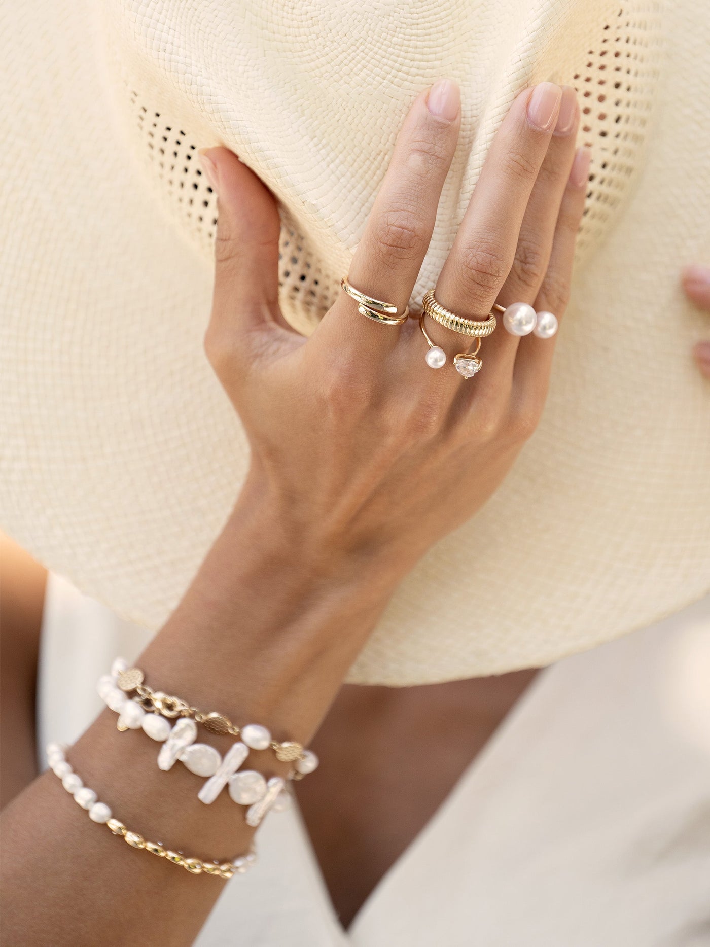 A hand adorned with multiple rings and bracelets elegantly holds a wide-brimmed straw hat, set against a light, neutral background suggesting a sunny outdoor environment.