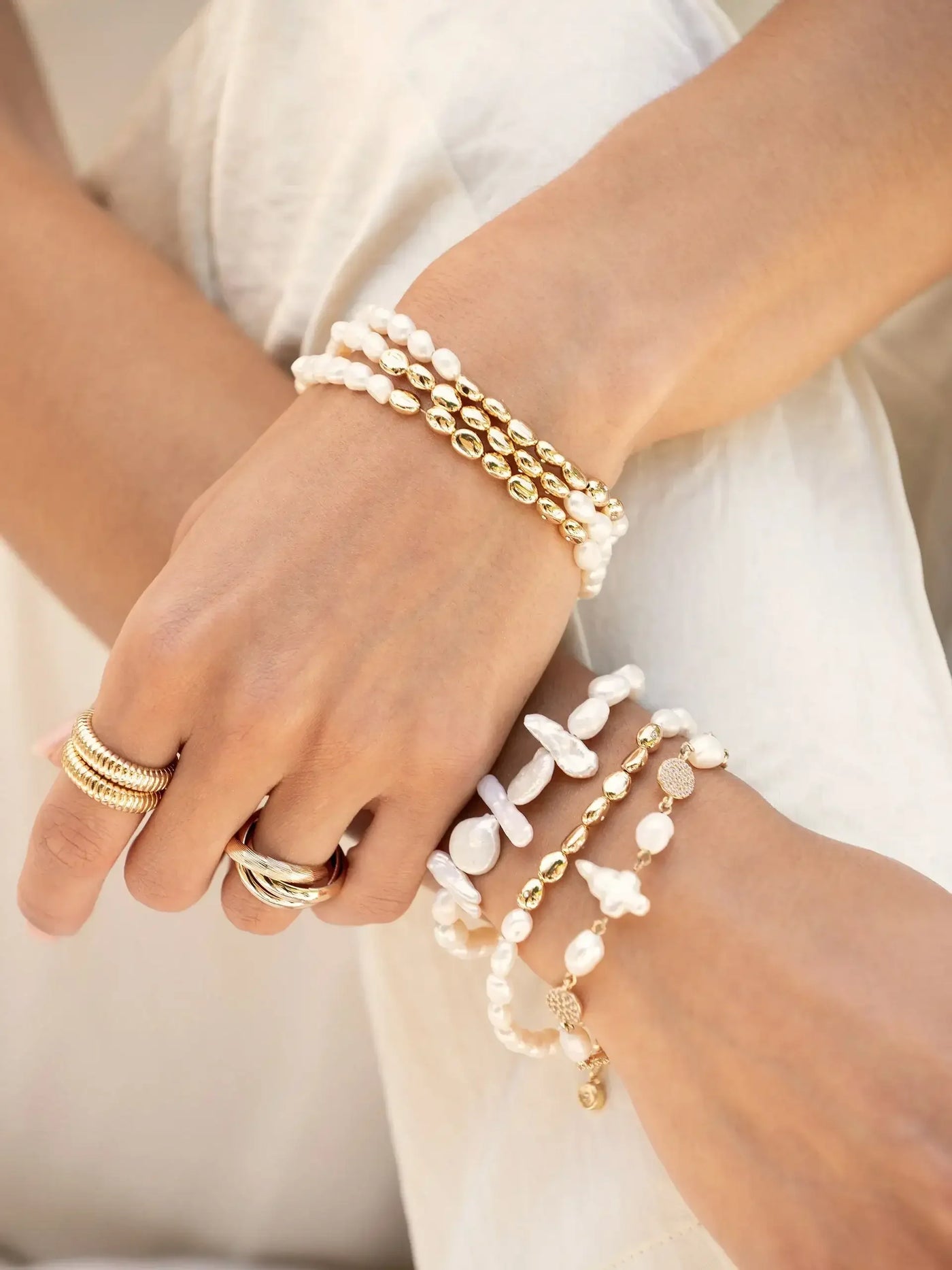 A pair of hands adorned with multiple bracelets, including gold and white pearl designs, gracefully rests against a soft, light-colored fabric background, showcasing a stylish accessory arrangement.