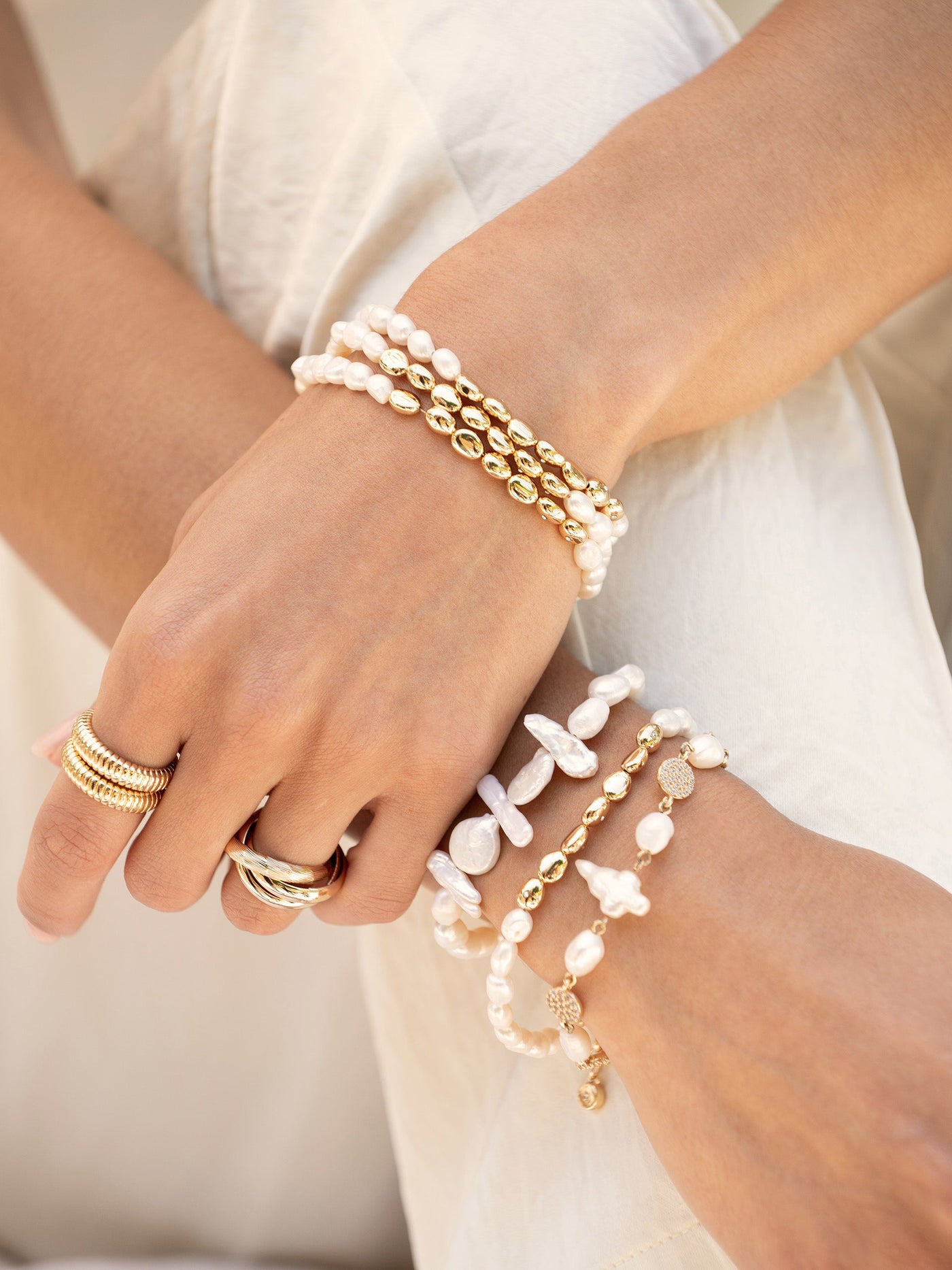 A pair of hands adorned with multiple bracelets, including gold and white pearl designs, gracefully rests against a soft, light-colored fabric background, showcasing a stylish accessory arrangement.