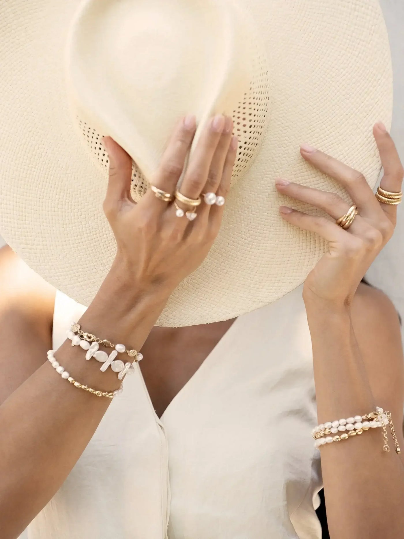 A hand, adorned with multiple rings and bracelets, holds a large, straw sunhat. The context suggests a sunny, fashionable setting, possibly outdoors, emphasizing a relaxed, stylish vibe.