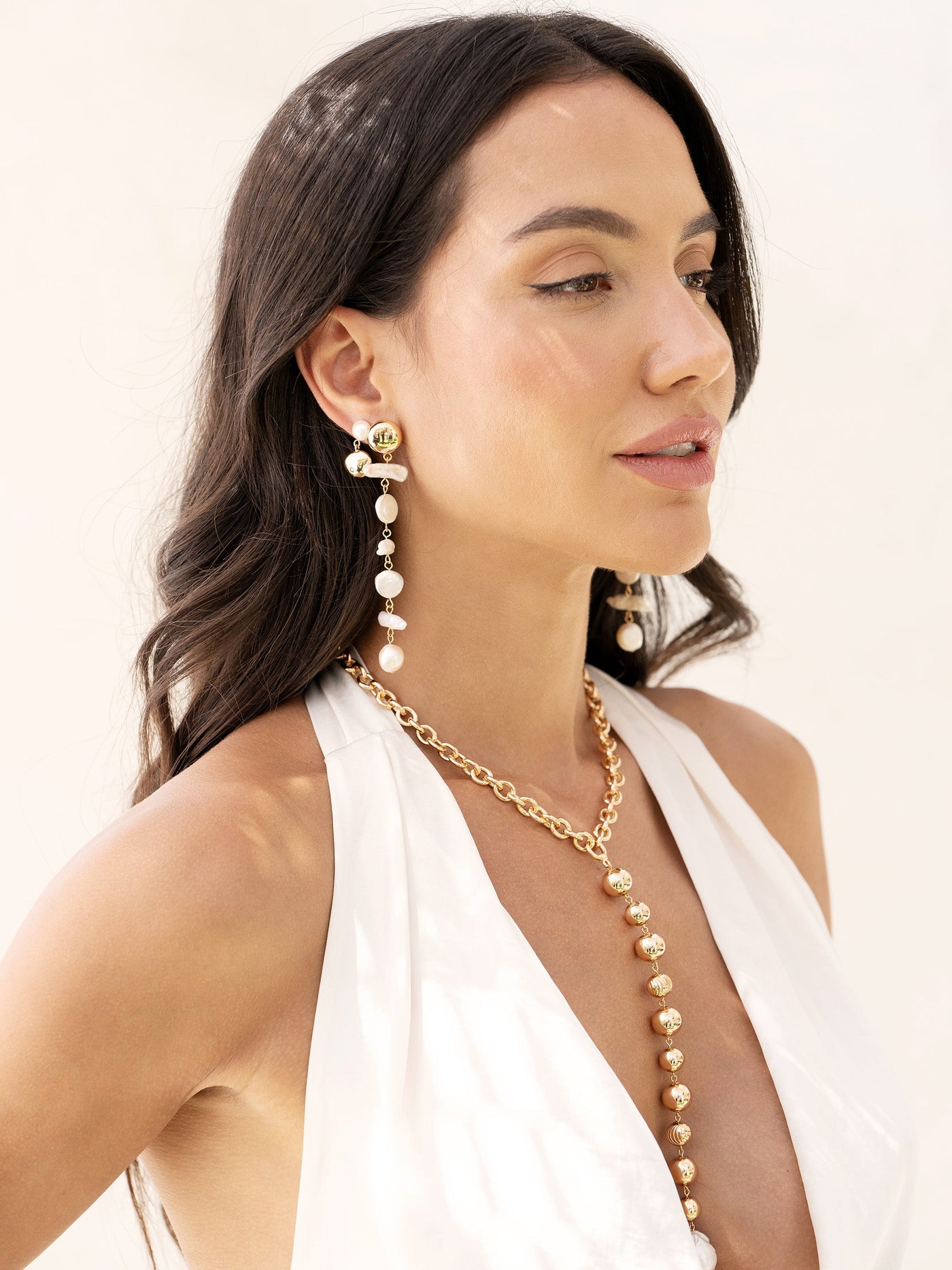 A woman wears bold pearl and gold earrings, with a matching gold necklace. She stands against a light, neutral background, showcasing her accessories and elegant attire.
