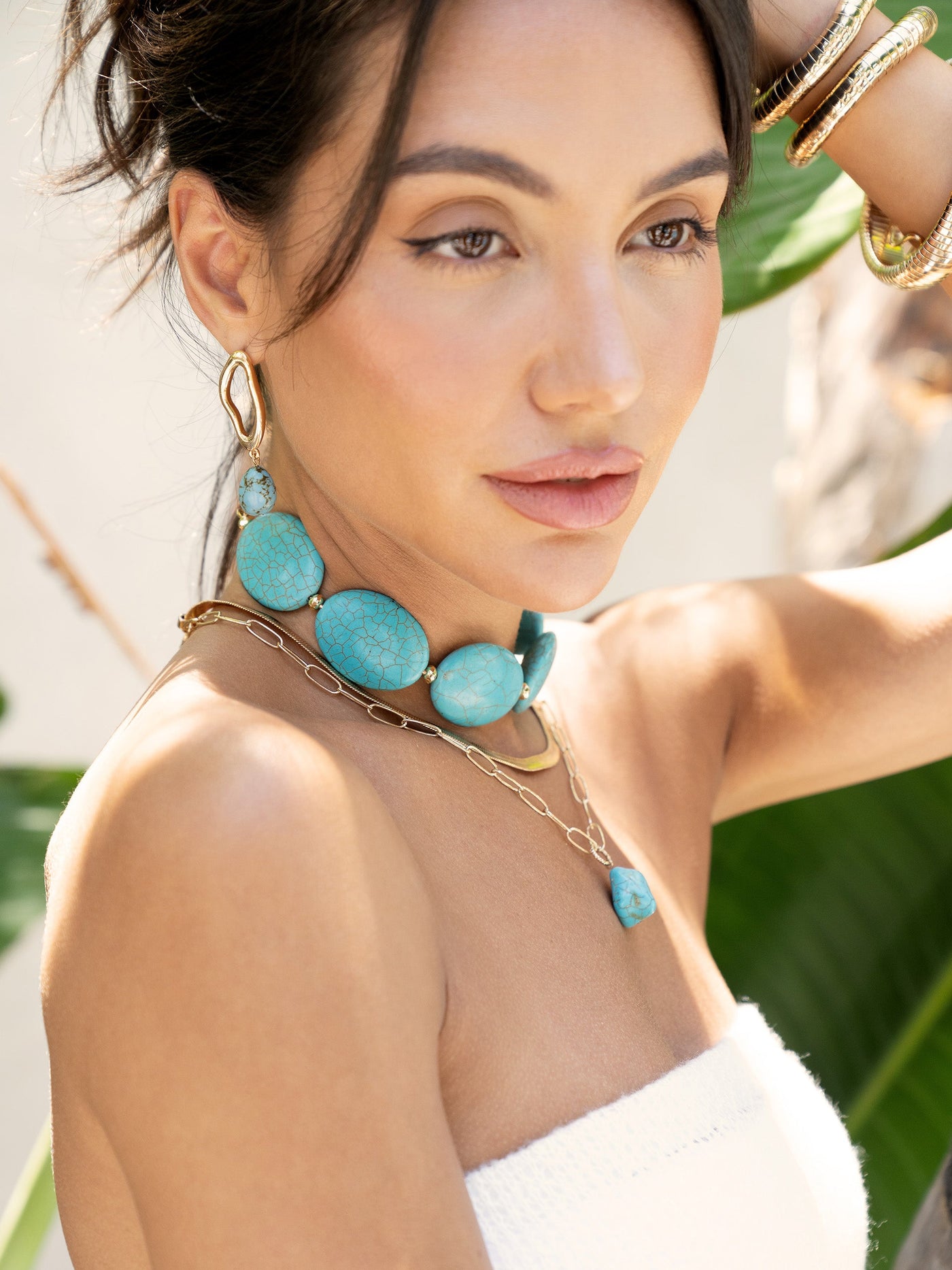 A woman poses elegantly, wearing turquoise jewelry and a strapless top. She gazes thoughtfully, with lush green foliage softly blurred in the background, enhancing the casual, sunny atmosphere.