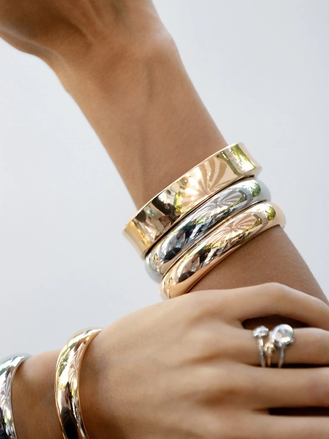 A hand adorned with multiple polished metallic bangles and a ring displays elegance. The setting is bright, with reflections suggesting an outdoor environment.