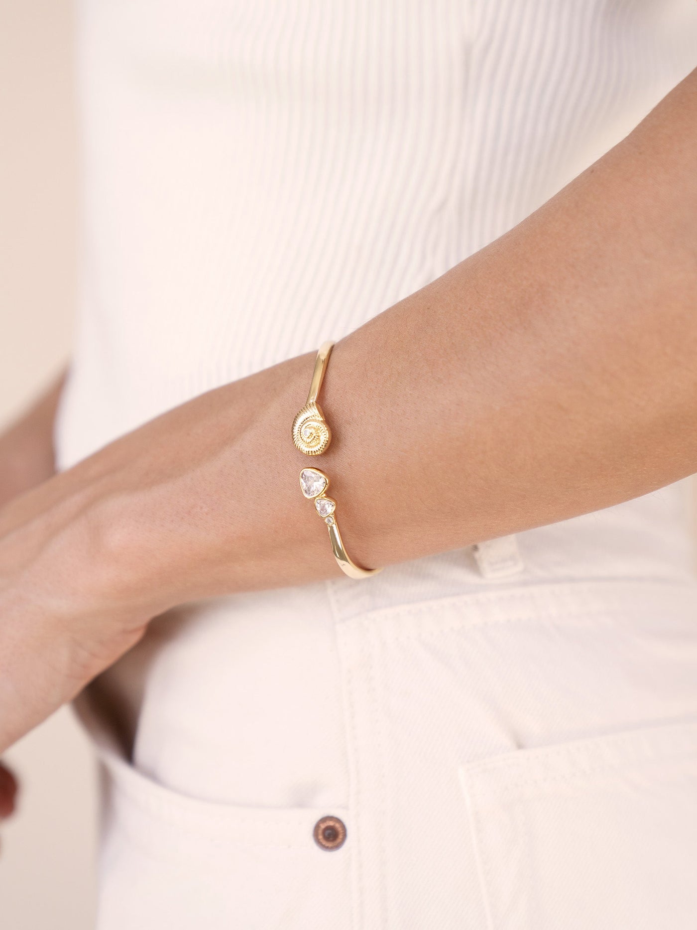 A gold bracelet with a spiral design and two small gemstones is worn on a wrist. The surrounding context features a light-colored, textured top and light-colored pants, suggesting a casual outfit.