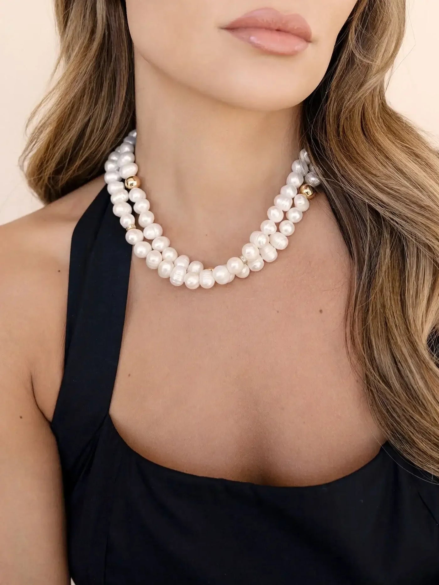 A two-stranded pearl necklace rests around the neck of a woman wearing a sleeveless, dark dress. Her lips are soft and neutral-toned, against a light background.