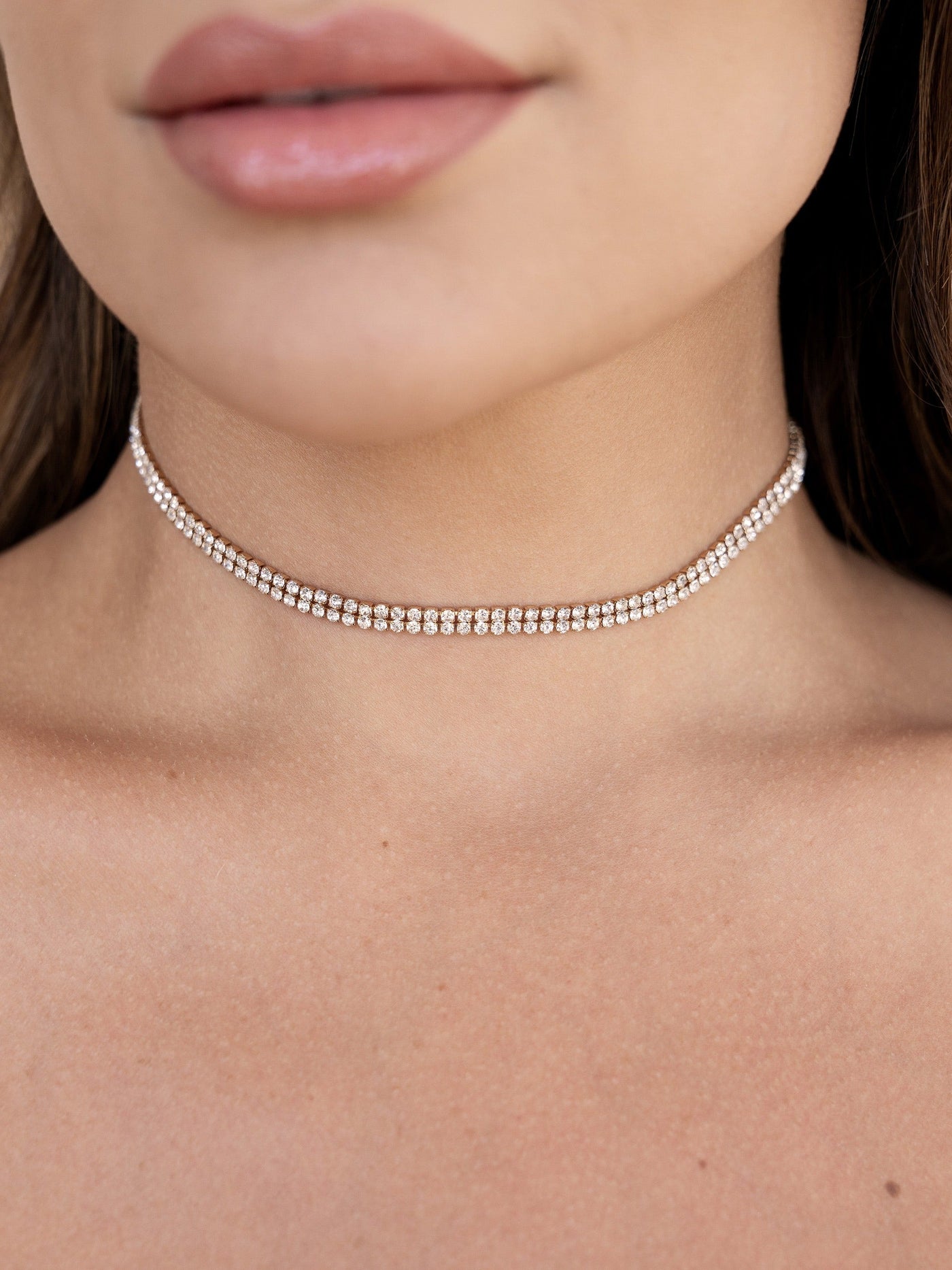 A close-up of a woman's neck showcases a sparkly choker made of small, shiny stones. Her lips appear soft and natural, against a smooth, lightly tanned skin background.