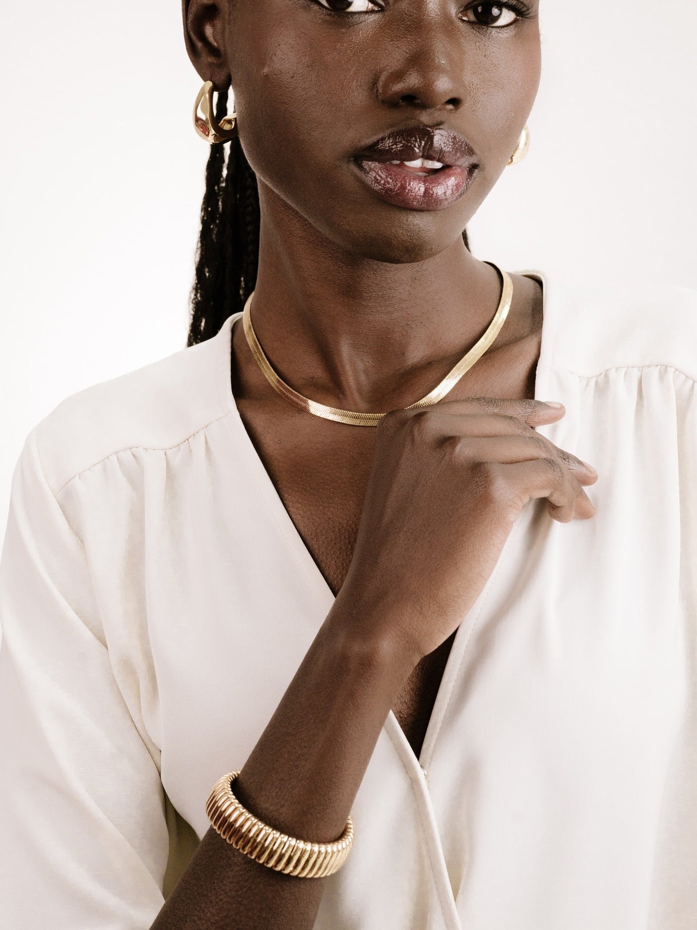 A person wears elegant gold jewelry, including hoop earrings, a choker, and a bracelet, while gently touching their neck and displaying a soft smile against a neutral background.