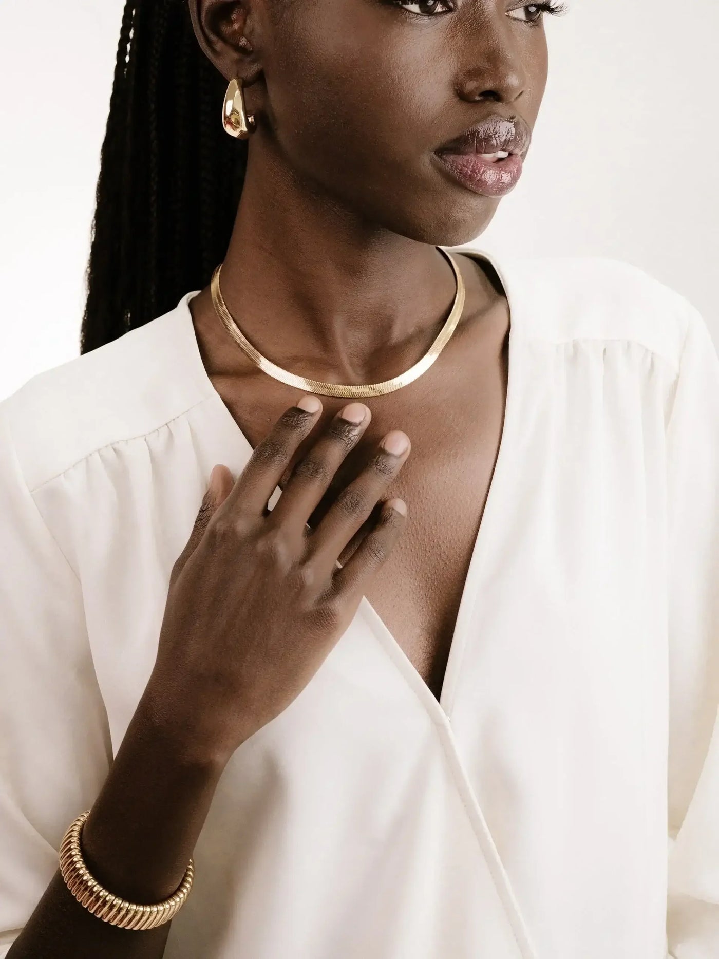 A woman in a white blouse touches her neck, adorned with a golden necklace and a matching bracelet, while wearing golden earrings. The background is neutral and soft, enhancing the jewelry's elegance.