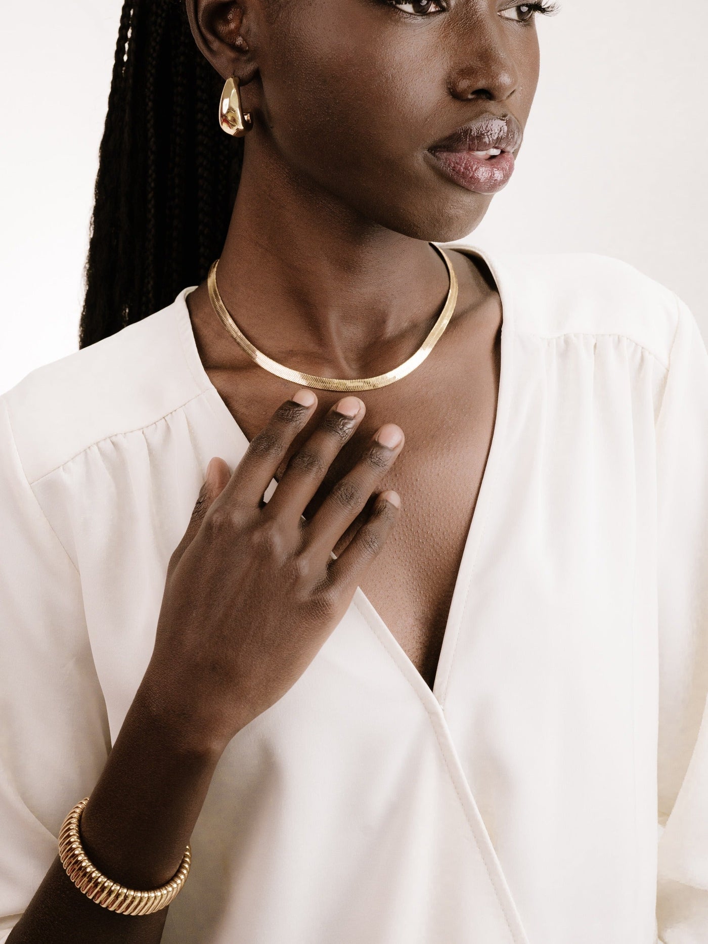 A woman in a white blouse touches her neck, adorned with a golden necklace and a matching bracelet, while wearing golden earrings. The background is neutral and soft, enhancing the jewelry's elegance.